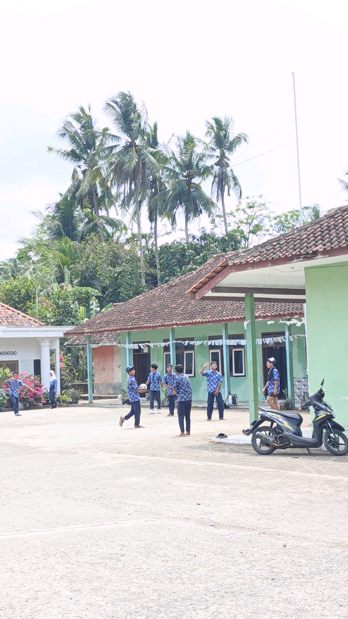 The image depicts an outdoor scene in Cibenda, Indonesia, featuring a group of individuals, primarily young men, gathered in what appears to be a schoolyard or community space. Several individuals are wearing matching blue and white patterned shirts and dark trousers, suggesting they might be students or part of a team. Some are actively engaged in a game, with one person holding a volleyball. Others are observing or standing nearby.
The environment includes two low-rise buildings with tiled roofs and verandas. One building is painted green, and the other is white. Palm trees and dense foliage surround the area, indicating a tropical climate. The ground is a paved, sandy surface, and a black and yellow scooter is parked to the right of the frame. The sky is overcast, suggesting a possibly humid day. The overall impression is of a casual, community-oriented activity taking place in a rural or semi-urban Indonesian setting.