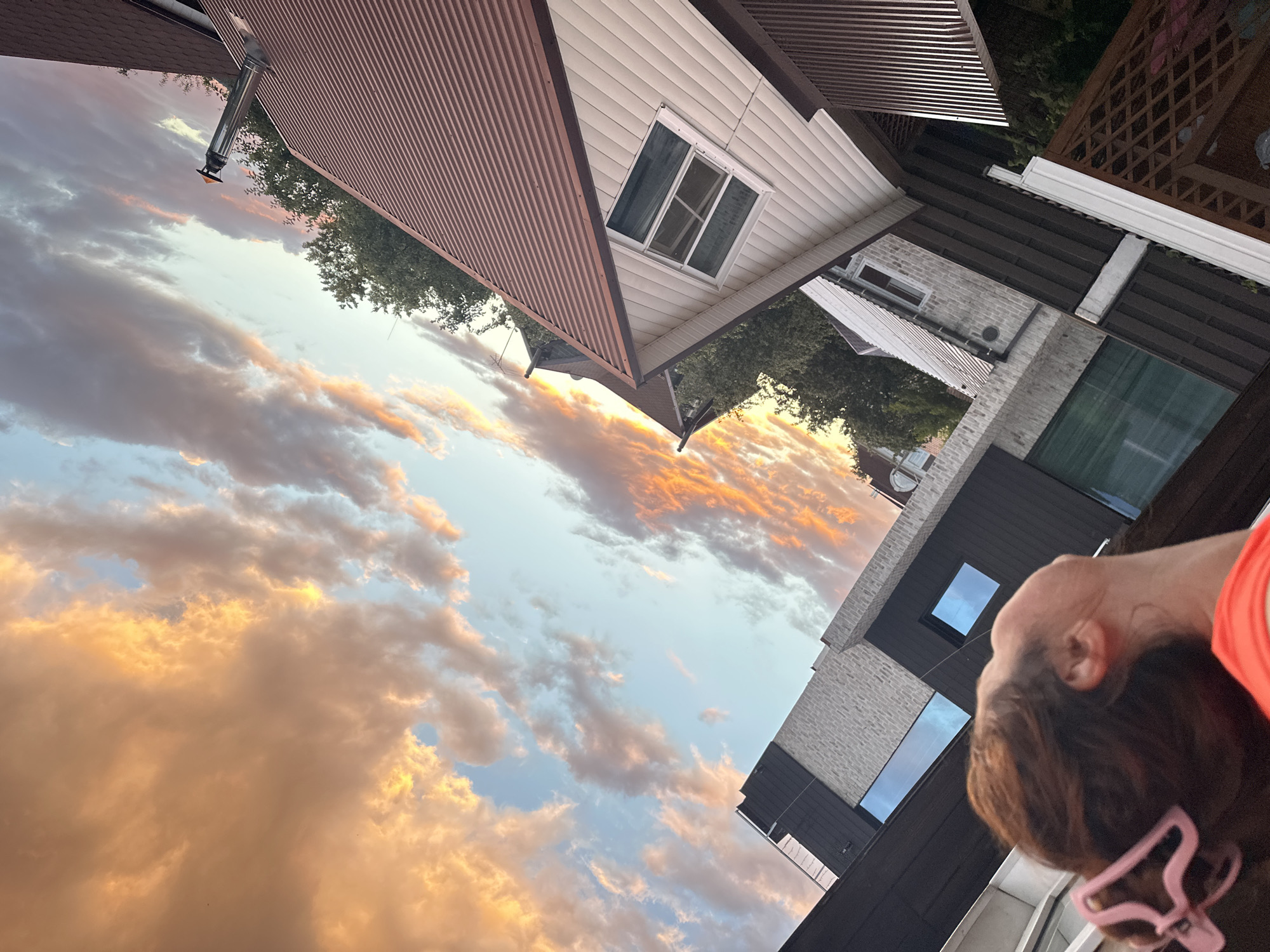 The image captures a view of the sky with dramatic clouds illuminated by sunset, seen from a residential area. The sky displays a mixture of orange, yellow, and blue hues. In the foreground, the back of a woman's head is visible, with her hair pulled back and secured by a pink clip. She is wearing an orange garment. The background features modern houses with pitched roofs, windows, and architectural details, suggesting an urban or suburban setting. Trees are also present, interspersed with the buildings. The image is oriented with the sky occupying the upper portion of the frame. The specific location in Nizhny Novgorod, Russia, cannot be determined from the visual information provided.