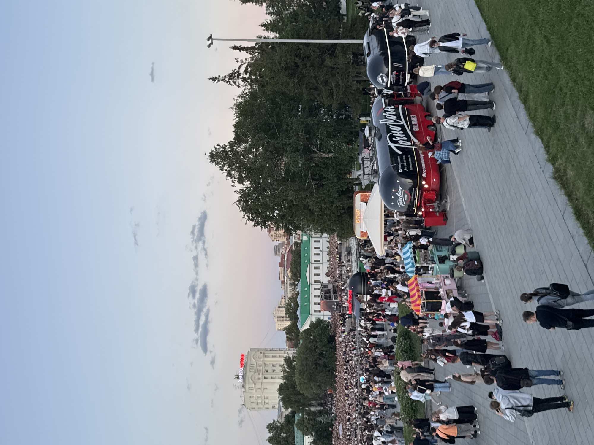 The image depicts a lively outdoor scene in Yekaterinburg, Russia, likely during an evening event. In the foreground, a paved walkway is populated by numerous people. Several food trucks are prominent, including two large black trucks with red accents labeled "Torno Corn GRILL MARKET" and another labeled "Spacebox." Other food stalls with colorful awnings are also visible. In the background, a dense crowd is gathered, suggesting a concert or festival. Beyond the crowd, a white building with red neon signage stands out, along with other urban structures and trees. The sky is a gradient of soft blues and pinks, indicating dusk.