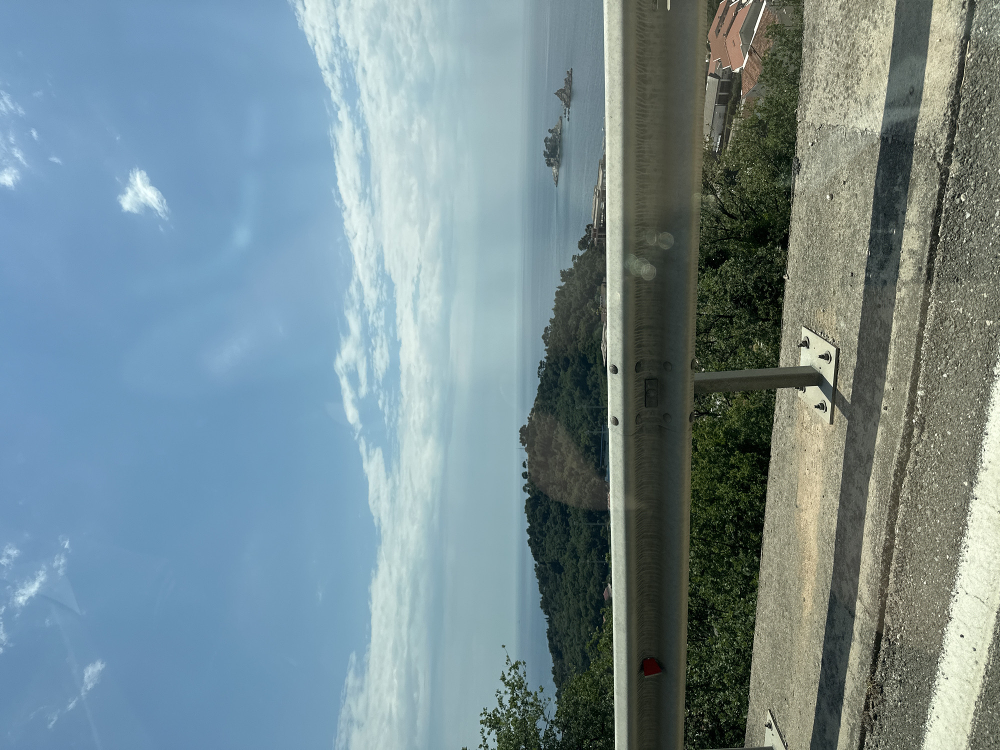 The image depicts a view from an elevated position in Brežine, Montenegro. A metal guardrail and concrete barrier dominate the foreground on the right side of the frame, bordering a road. Beyond the guardrail is a steep, densely forested slope leading down towards a body of water. In the distance, small rocky islands dot the sea. The sky is blue with scattered white clouds. A few houses with red tile roofs are visible amidst the trees on the right.