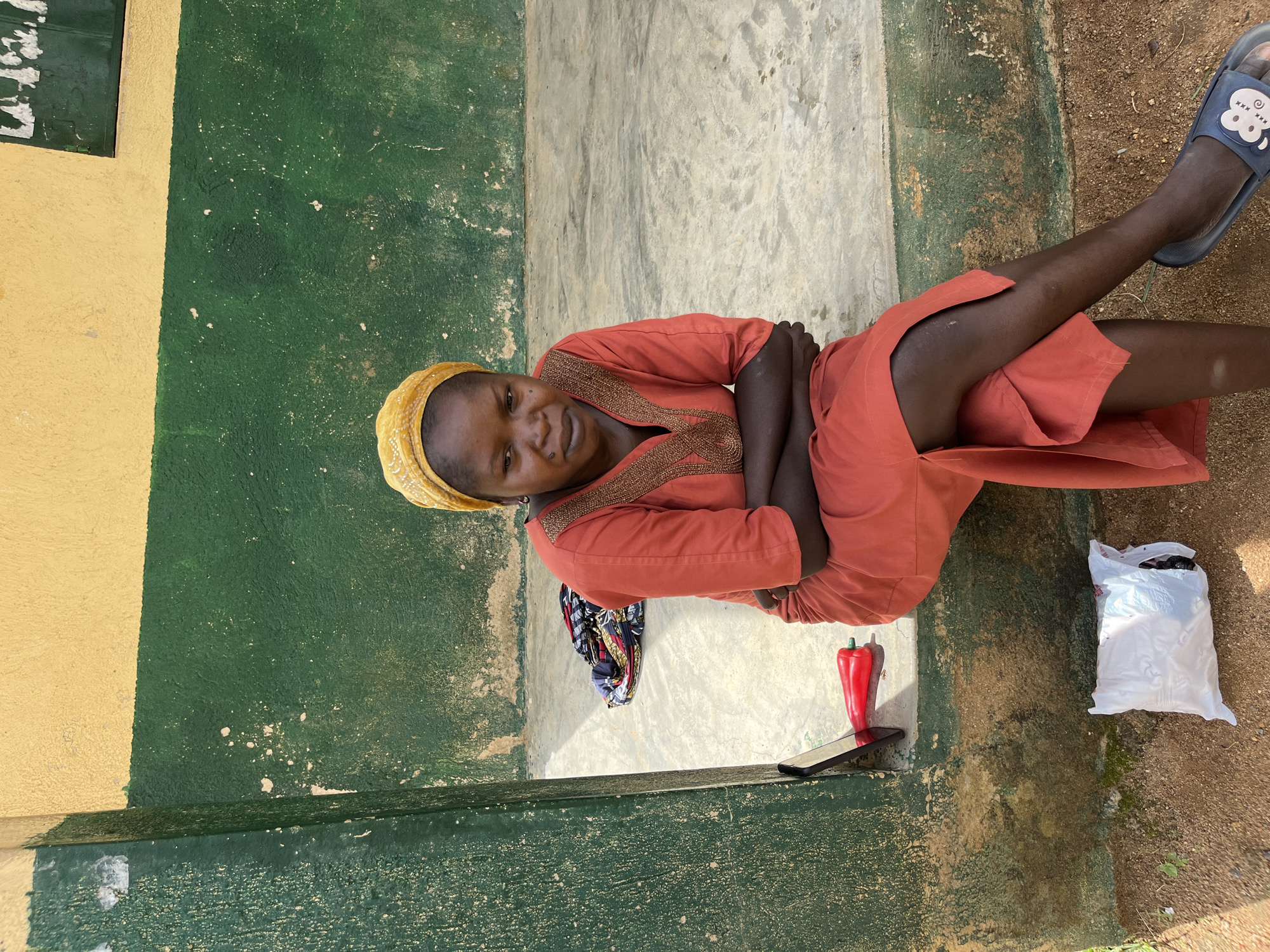 A Black female sits cross-armed on a concrete ledge against a two-toned yellow and green wall in Bukuru, Nigeria. She wears a vibrant orange dress and a yellow headscarf. Her arms are crossed, and her legs are crossed at the knee. To her left, on the concrete, are a red pepper-shaped object, a tablet, and a folded bundle of patterned fabric. On the ground to her right is a white plastic bag. The wall is textured and appears weathered, with white splotches visible on the green section. The ground is a mixture of dirt and sand.