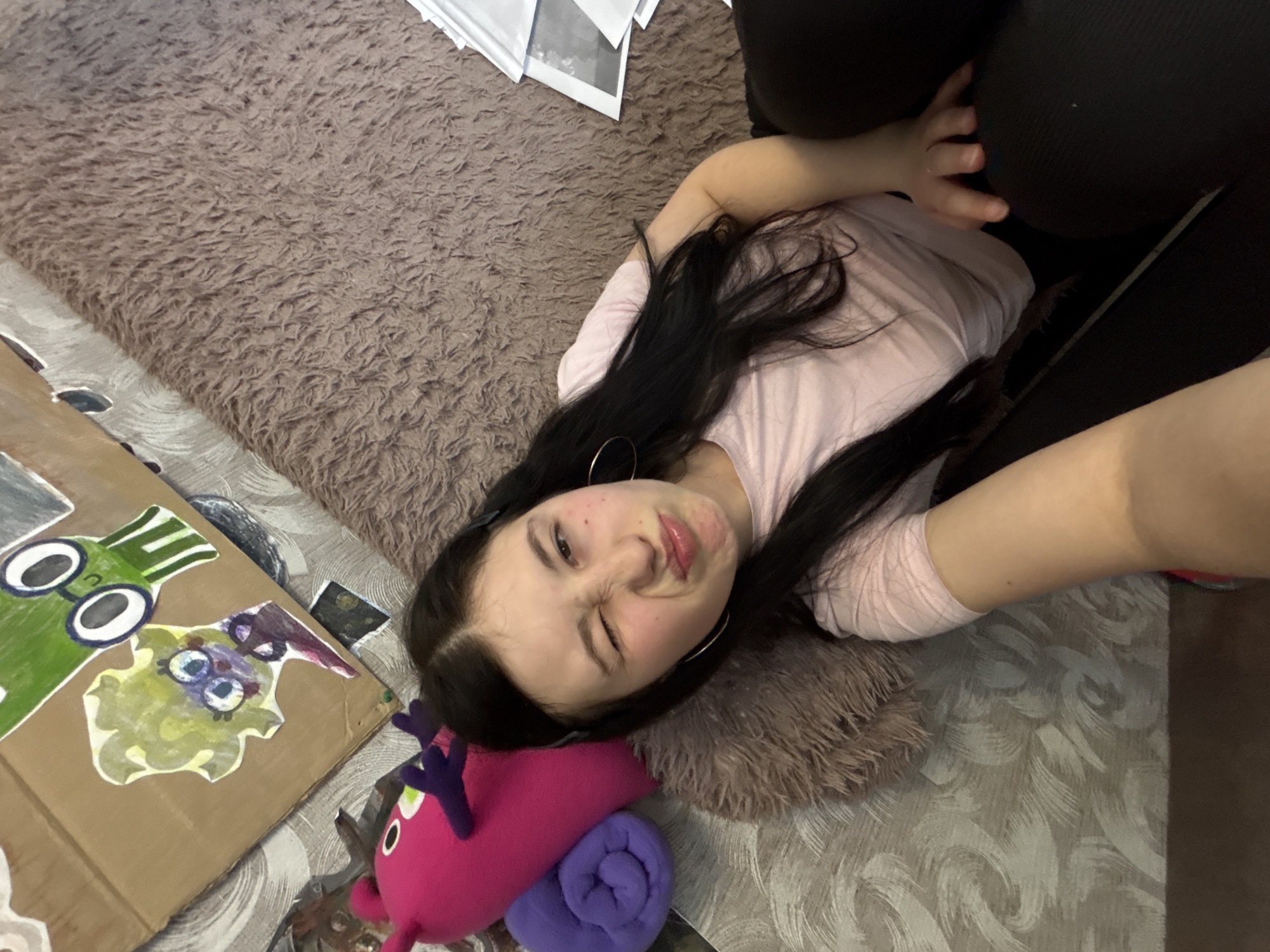 A young woman with long, dark hair is lying on a fluffy brown rug. She is wearing a light pink long-sleeved shirt and dark leggings. Her head is resting on a pink and purple plush toy, next to a cardboard box decorated with colorful drawings. The woman is winking at the camera. The background includes a marble-patterned surface and papers. This photo was taken in the city of Фоки, Russia.