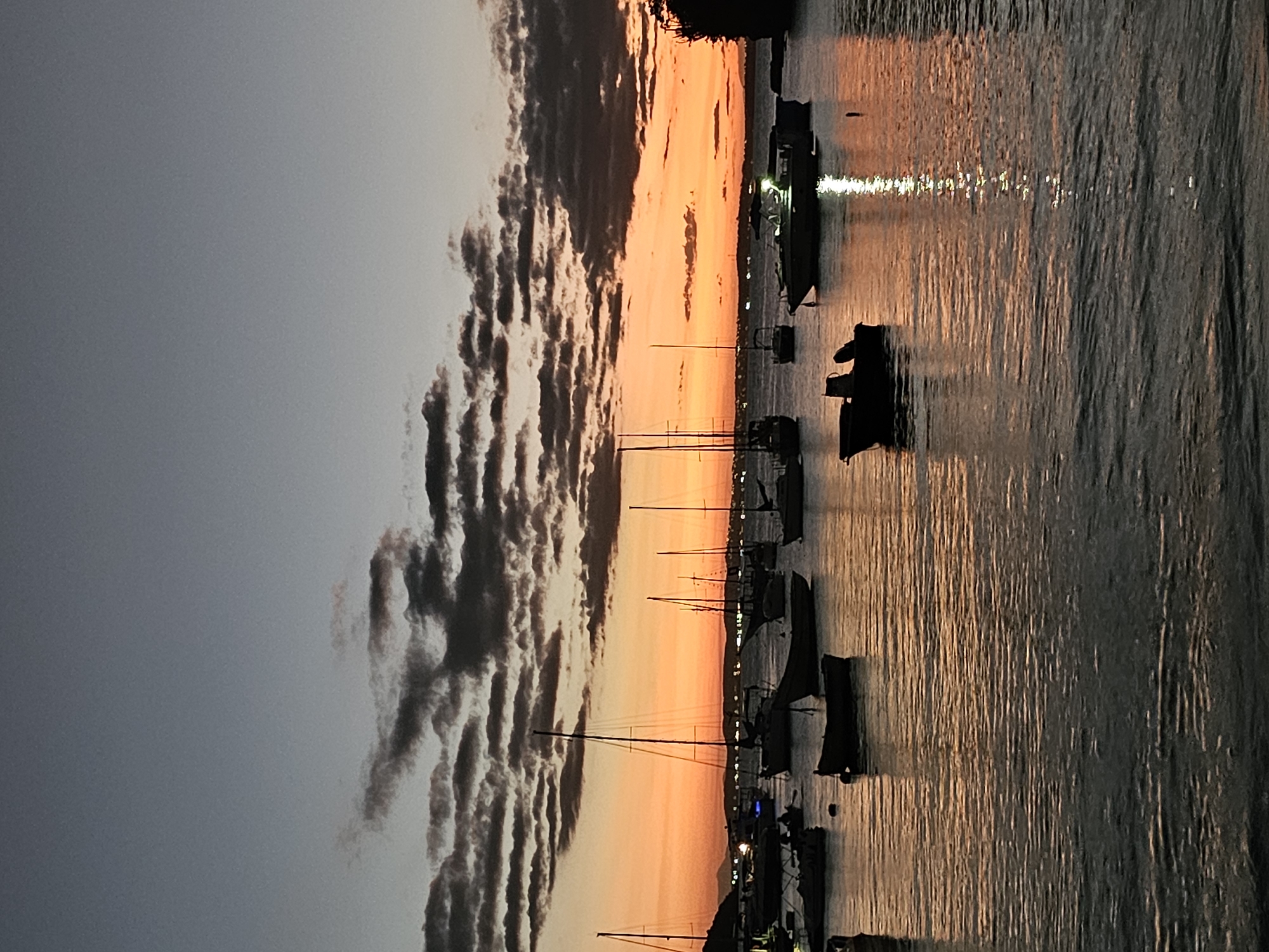 The image depicts a harbor scene in Armação Dos Búzios, Brazil, during a vibrant sunset. Silhouetted sailboats and other small watercraft dot the calm water, their masts pointing towards the orange and yellow sky. The water reflects the colorful sky. Cumulus clouds are on the left side.