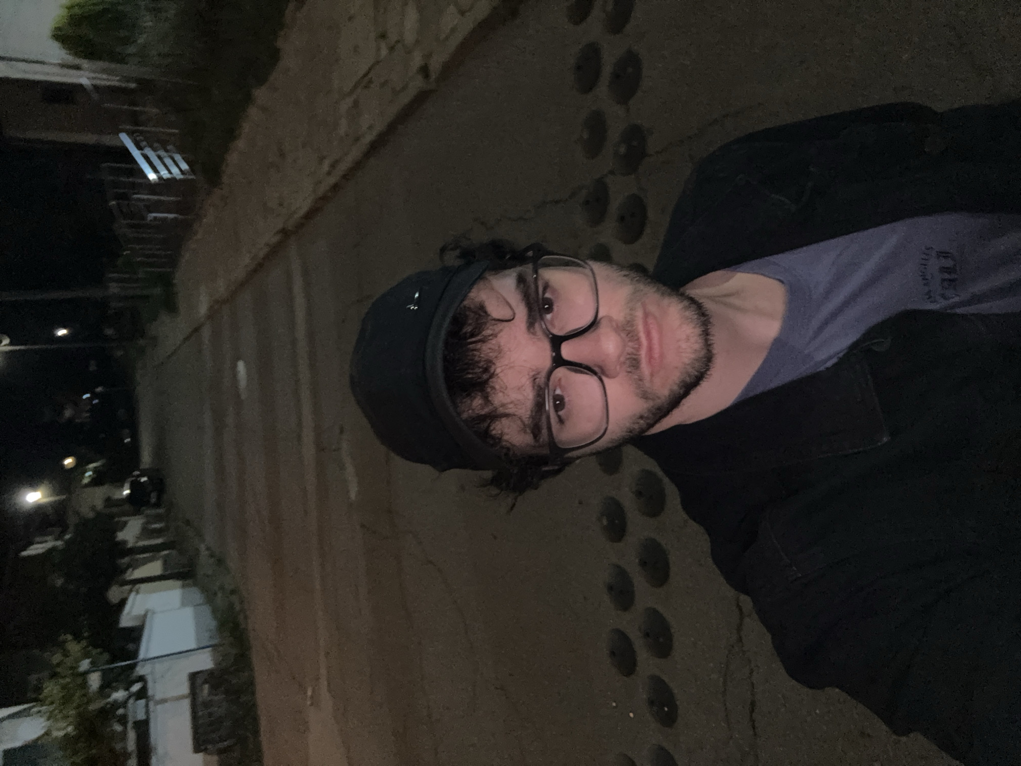 A young man with dark curly hair, wearing glasses and a black baseball cap, stands in the middle of a street at night. He is looking directly at the camera and is dressed in a dark jacket over a blue t-shirt. The street appears to be paved and has speed bumps. Buildings and trees are visible in the background, illuminated by streetlights. The setting is consistent with a residential area in Blagoevgrad, Bulgaria, possibly a side street or a quiet road.