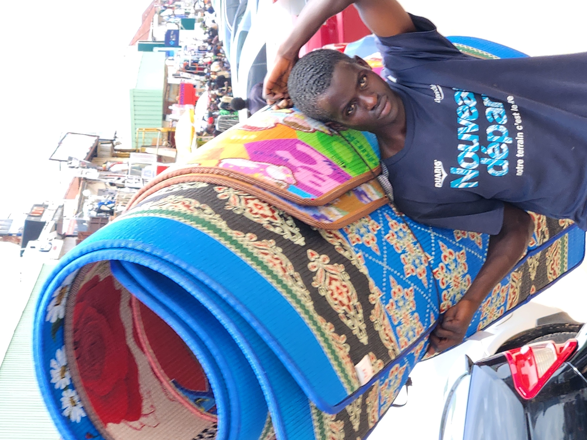 A dark-skinned young man carries a large, rolled-up mat over his right shoulder. The mat is brightly colored with a blue border, a repeating red and yellow floral pattern on a black background, and a blue section with a diamond pattern. The man is wearing a dark blue t-shirt with white text that reads "Nouveau départ notre terrain c'est le re". He is looking directly at the camera, and his left hand is resting on the mat. In the background, there are blurred figures of people, cars, and buildings, suggesting an outdoor market or street scene. The sky is bright and appears to be day time.
