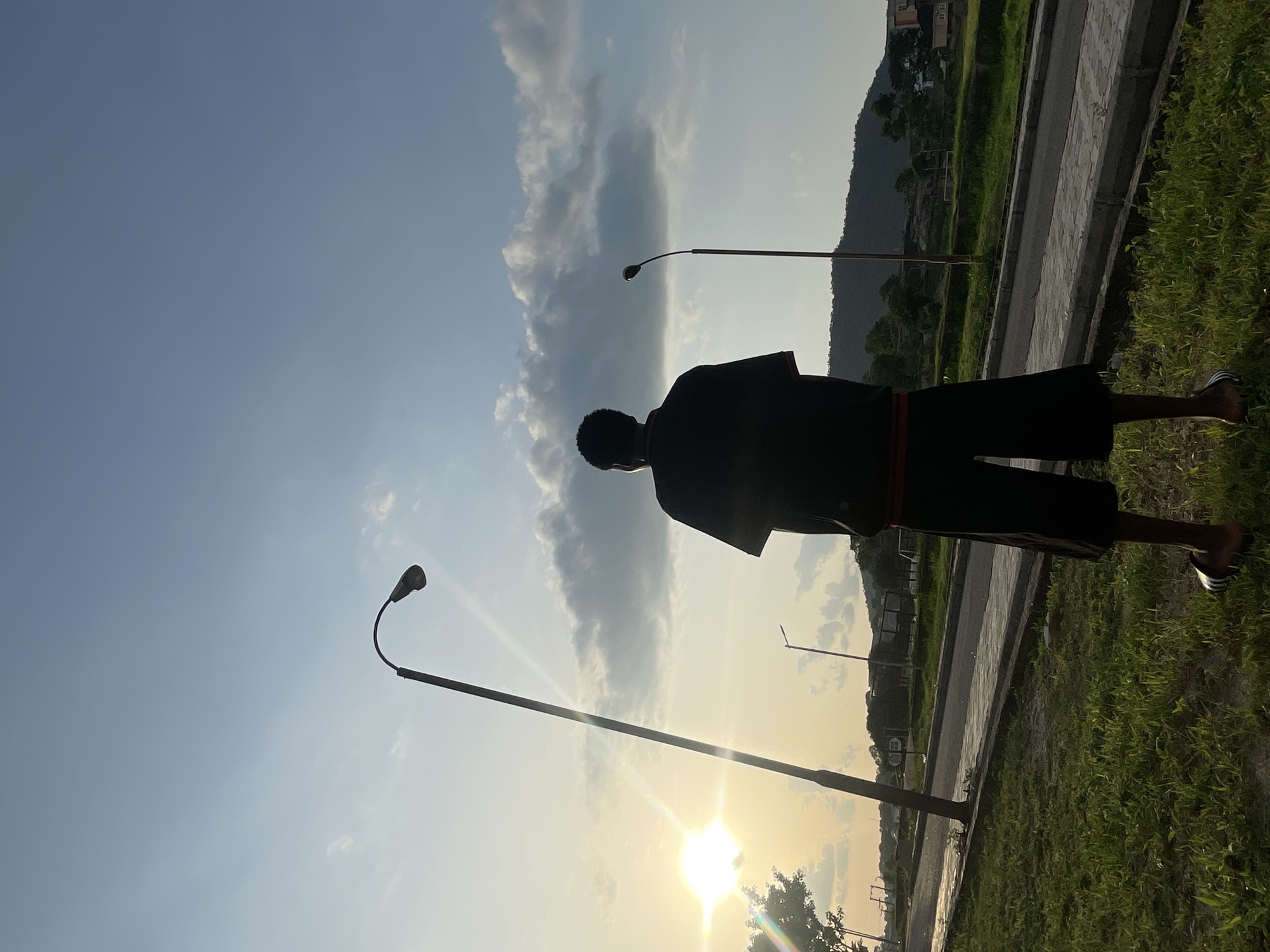 A person stands on a grassy field in Lokoja, Nigeria, their body silhouetted against a bright, setting sun. They are wearing dark clothing and sandals. Several streetlights are visible, lining a paved walkway that borders the field. In the background, there is a mountain. The sky is partly cloudy.