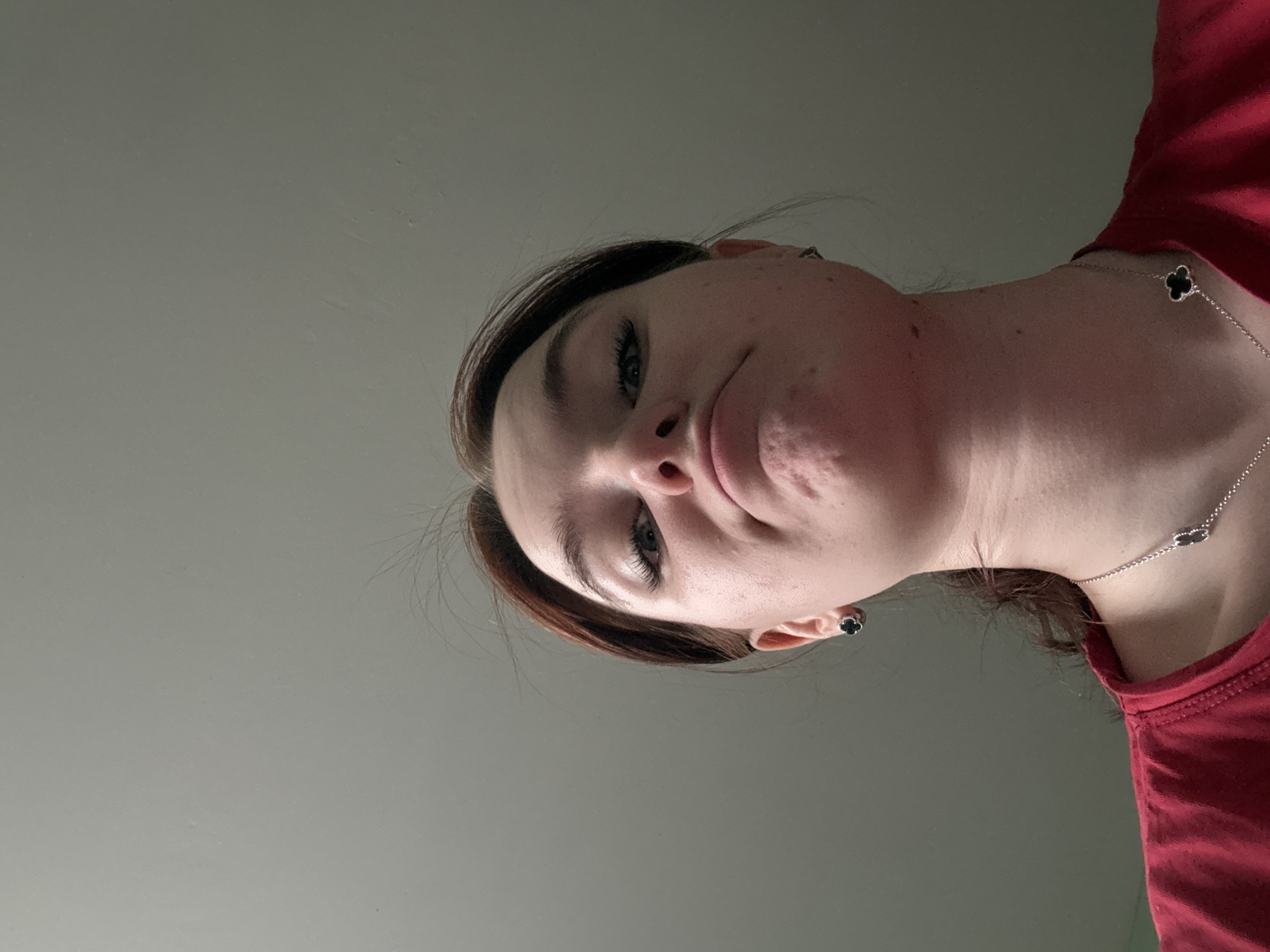A low-angle close-up shot of a woman. She has fair skin, dark blonde hair, and is wearing a red top and a silver necklace with clover-shaped pendants. She has dark earrings. The background is a plain, light gray color. The location is Unknown, Russia.