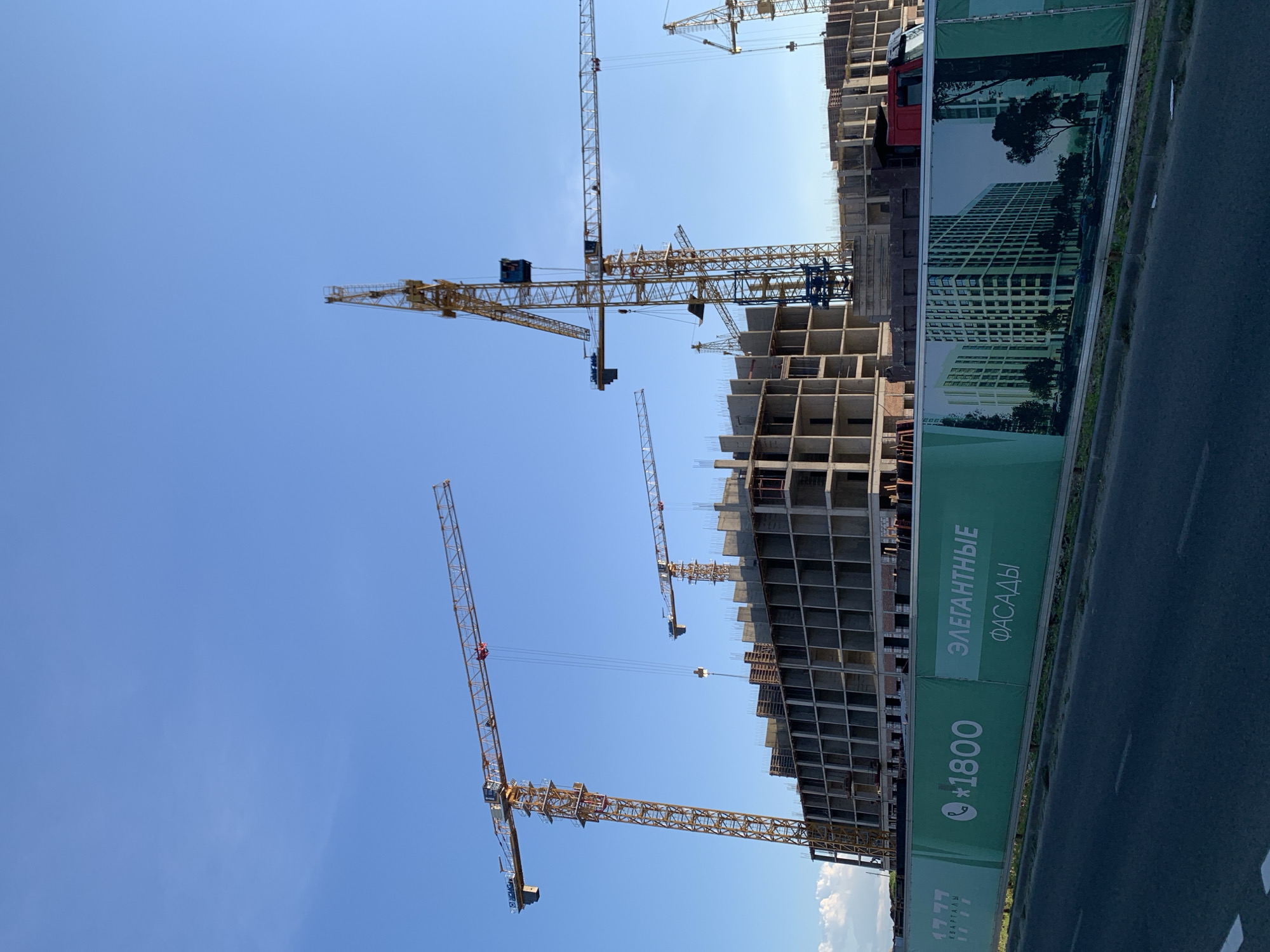 The image captures a construction site under a clear, bright blue sky, suggesting a sunny daytime. The primary subjects are multiple tall construction cranes, their yellow metal structures stark against the sky. One crane dominates the foreground, its long boom extending horizontally. In the background, a multi-story building is under construction, with its concrete framework exposed, showing multiple floors and structural elements.
To the right of the construction site, a green banner is stretched along a fence. This banner features text and an image. The text is in Russian and includes a phone number, "*1800", and phrases like "ЭЛЕГАНТНЫЕ ФАСАДЫ" (Elegant Facades) and "КВАРТАЛЫ" (Quarters). The banner also displays a printed image of a completed apartment building, suggesting the project's intended outcome. A red truck is partially visible behind the banner and next to the building. The overall scene conveys a sense of ongoing development and progress in an urban or suburban setting. The clear sky indicates fair weather.