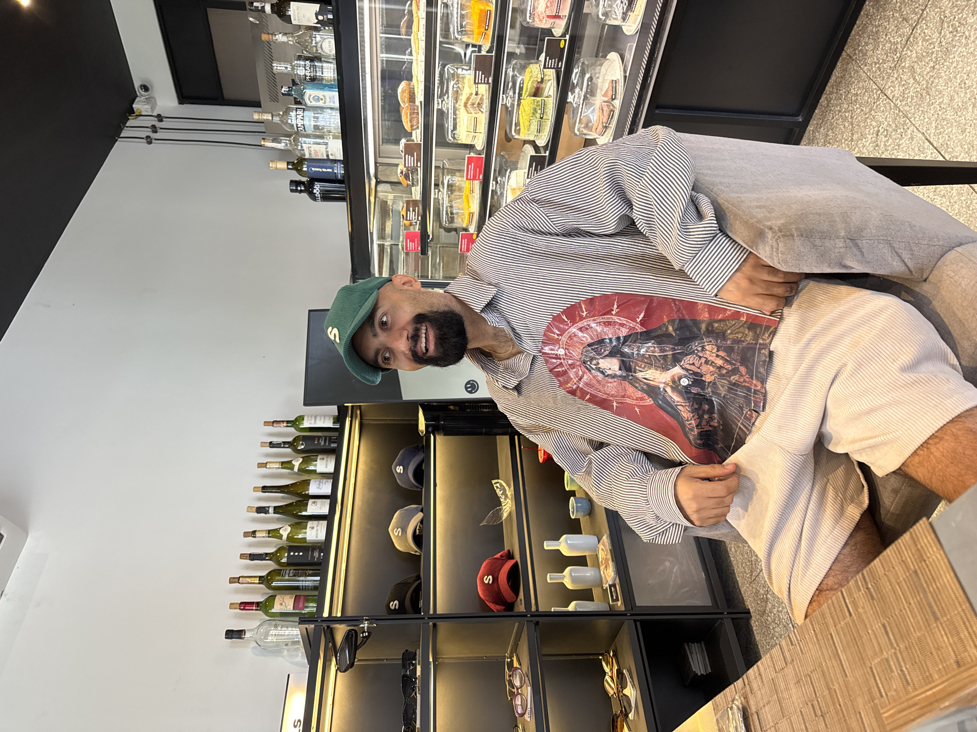 A medium shot shows a man sitting in a chair in what appears to be a retail establishment or cafe, with a display of cakes and bottles in the background. He is wearing a green baseball cap, a horizontally striped button-down shirt with a large graphic print on the front, and beige shorts. The man is looking off to the right and smiling. The background displays shelves stocked with various bottles of wine and liquor, as well as a refrigerated display case filled with cakes and pastries. Another shelf holds baseball caps. The flooring is tiled, and the walls are white and black. The image does not provide specific information to locate it in Ko Samui District, Thailand.