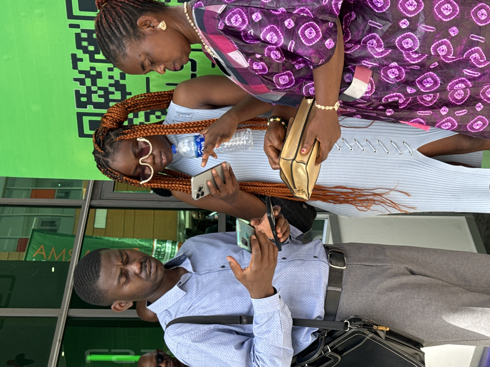 A man wearing a light blue shirt, gray pants, and a black bag is holding a cell phone in his right hand. A woman in a purple and white patterned dress and a woman in a light blue dress are standing behind him. The woman in the light blue dress is holding a cell phone in her left hand and a water bottle in her right hand. The woman in the purple dress is holding a gold purse in her right hand. A QR code and the word "AMS" are visible in the background. The location is Alaguntan, Nigeria.