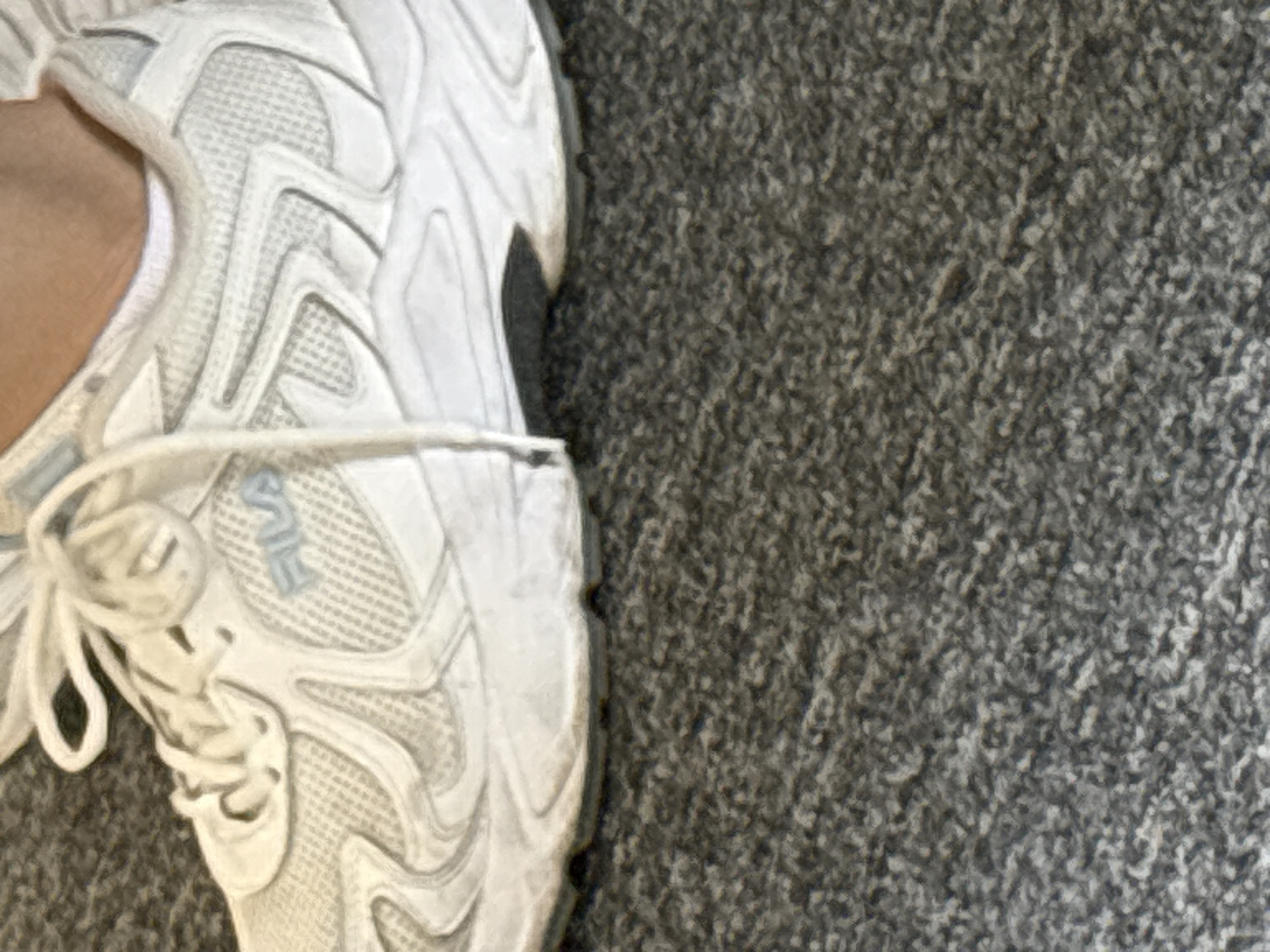 The image shows a close-up, angled view of a white Fila sneaker resting on a gray, textured carpet. The sneaker is predominantly white with subtle gray accents and a blue Fila logo on the side. The laces are white and tied loosely. The sole of the shoe is white with a darker gray strip along the bottom edge. The carpet appears to be indoor carpeting. There is no discernible indication of the shoe's location within a city or specific environment.