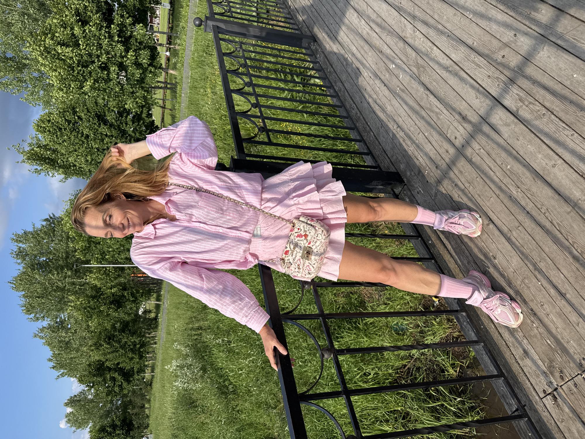 A woman stands on a wooden bridge in Kirishi, Russia. She is wearing a pink striped button-down shirt, a pink ruffled skirt, pink socks, and pink athletic shoes. She is carrying a small, patterned handbag. The woman is holding her hair with her right hand and leaning on a black metal railing. Behind her, there is a grassy area with trees and a bright blue sky. The lighting suggests it is daytime, likely late afternoon due to the shadows cast on the bridge.