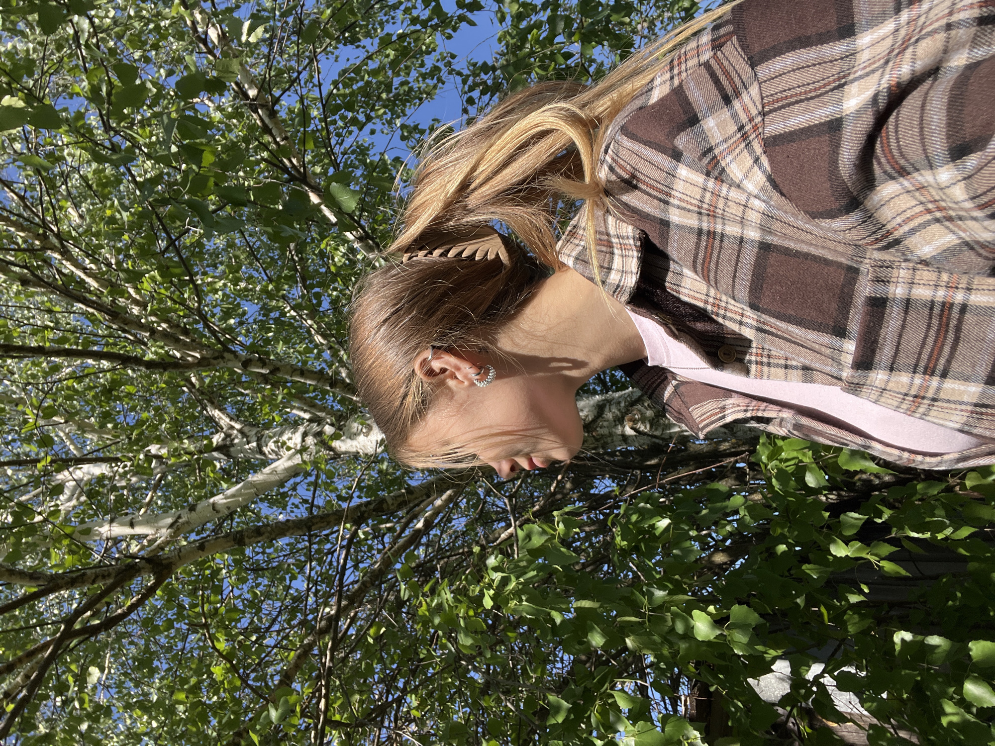 A woman stands under a tree on a sunny day. The tree has green leaves and white bark, suggesting it could be a birch tree, commonly found in Russia. The woman is wearing a brown and tan plaid shirt over a pink top. Her blonde hair is partially pulled back with a brown hair clip. She is looking slightly down and away from the camera. The background shows a clear blue sky. The location is specified as Фоки, Russia.