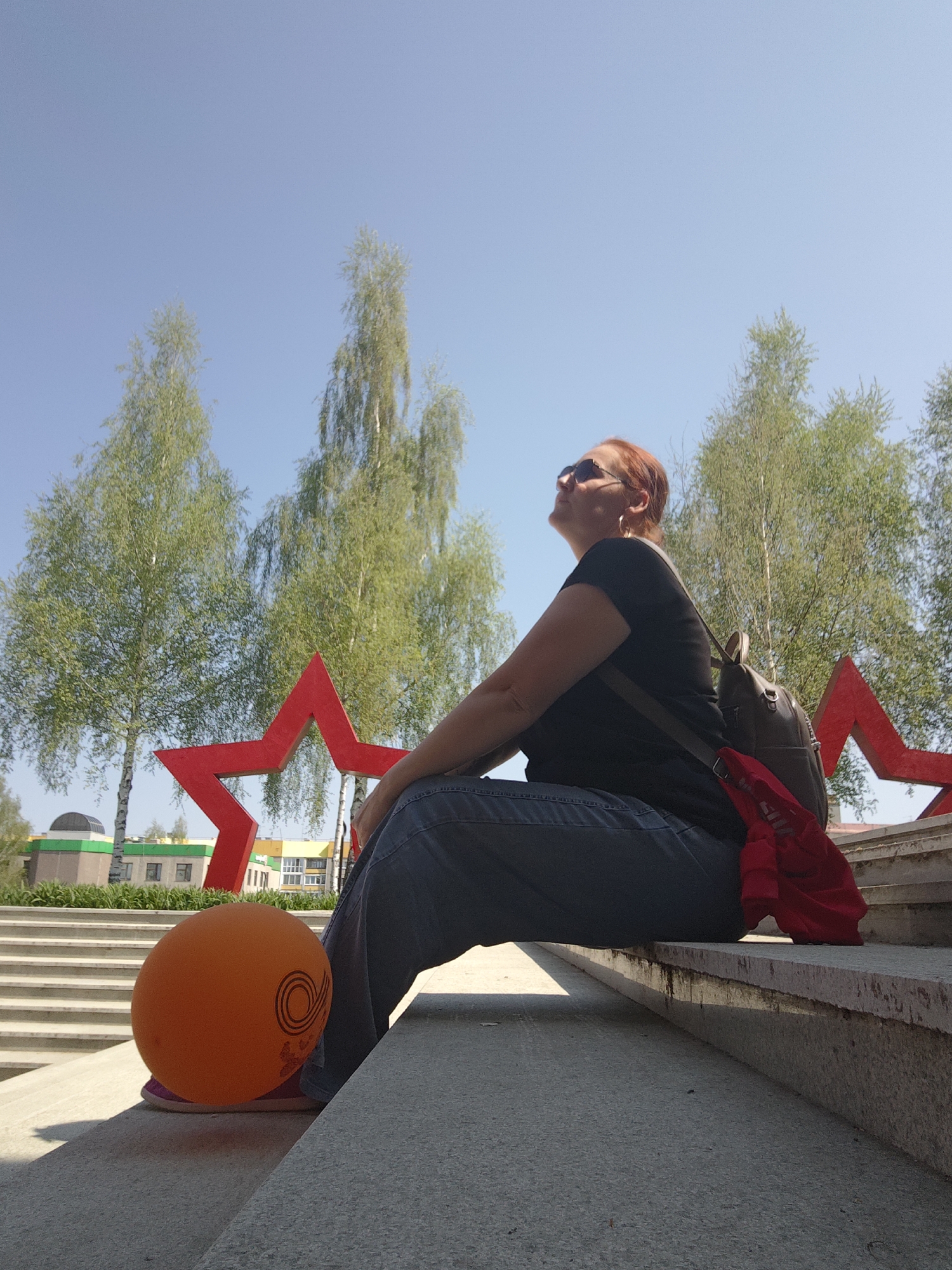 A woman with red hair and sunglasses is sitting on concrete steps. She is wearing a black t-shirt and blue jeans. She has a backpack on her back. An orange ball is next to her foot. Behind her are green trees and two red stars. In the background, buildings are visible. The location is Марковский, Russia.