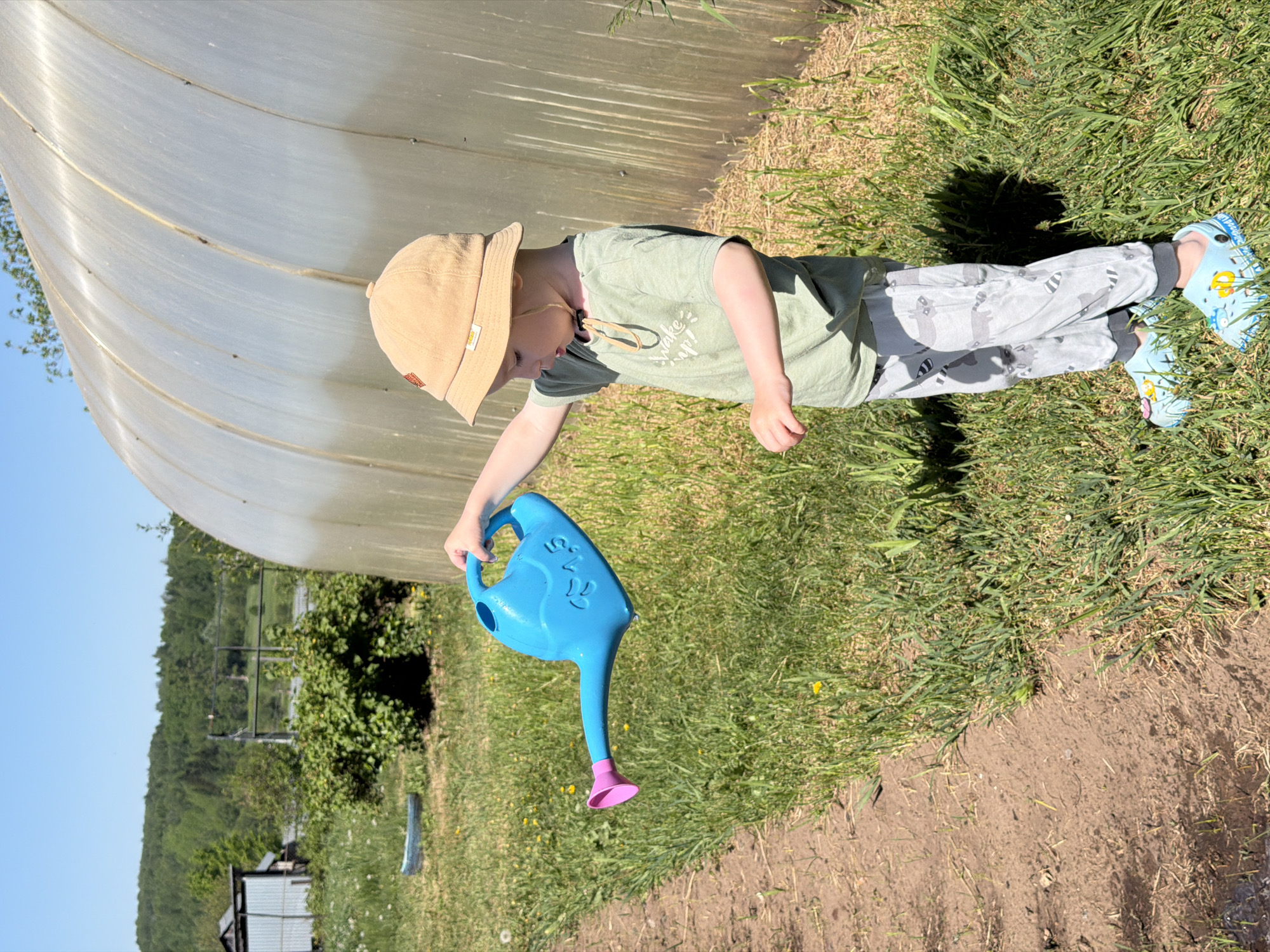 A child in Уральское, Russia, wearing a beige hat, green t-shirt, patterned grey pants, and blue Crocs stands on grass next to a low tunnel greenhouse. The child holds a blue watering can with a pink nozzle. The background shows trees, grass, a building, and a blue sky.