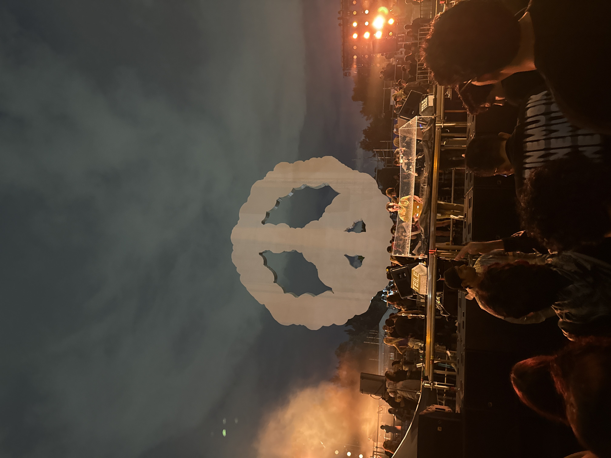 A wide-angle, eye-level outdoor shot captures a nighttime concert scene at the Autodromo Hermanos Rodríguez in Mexico City, Mexico. The focal point is a massive, stylized white peace sign sculpture dominating the upper center of the frame, silhouetted against a dark, cloudy sky. Below the sculpture, a crowd of concert-goers fills the lower portion of the image, their faces largely obscured or out of focus. On a raised stage in the middle ground, a DJ in a yellow shirt is visible, surrounded by equipment and illuminated by stage lights. To the right, a wall of bright orange lights illuminates the sky, casting an eerie glow. Smoke or fog machines are active on the left side of the stage, creating a hazy orange cloud that partially obscures the crowd and the peace sign. The overall atmosphere is one of excitement and energy, typical of a large outdoor music festival.