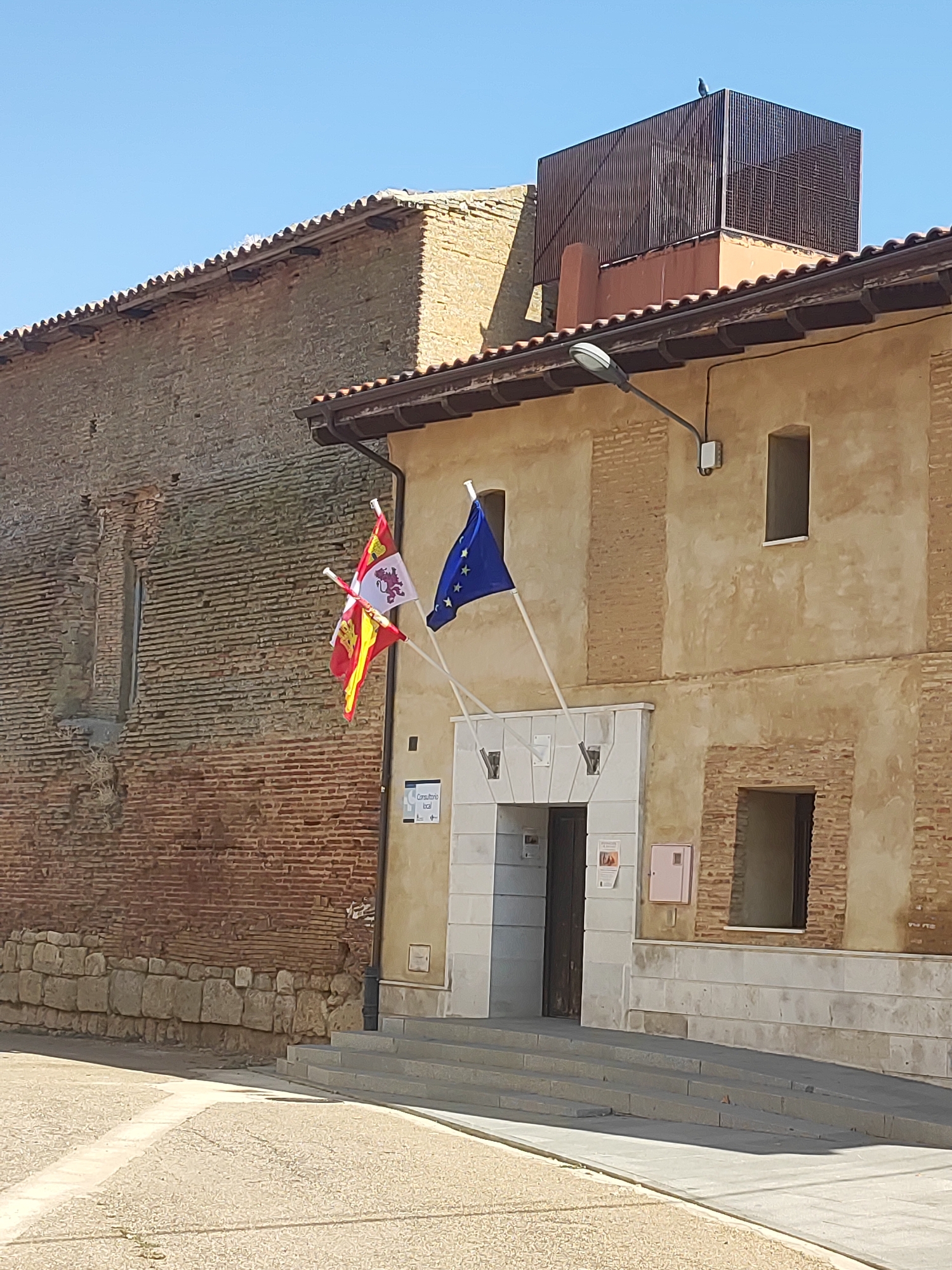 The image shows the exterior of a building in Autillo de Campos, Spain, under a clear blue sky. The building has two distinct sections: a foreground building with a light-colored, stuccoed facade and a recessed entrance with stairs leading up to it, and a background building constructed with rough-hewn bricks.

On the facade of the foreground building, two flags are visible, mounted on poles. The flag on the left is red and yellow, with a white crest featuring a lion. The flag on the right is the European Union flag, blue with yellow stars. A streetlamp extends from the building above and to the left of the flags. There are several windows on the building's facade, some rectangular and some with barred openings. A sign with the text "Consultorio local" is affixed to the wall near the entrance.

The background building is characterized by its exposed brickwork, appearing older and more weathered than the foreground structure. The brick wall has a rough texture and is punctuated by a single, tall, narrow window. At the top of the background building, a dark, metal structure is visible against the sky, with a bird perched on its surface.

The ground in front of the building is paved with light-colored cobblestones, with a defined pathway marked by a lighter strip of paving. Shadows are cast on the ground, indicating sunlight from the right. The overall scene suggests a public or administrative building in a town setting.