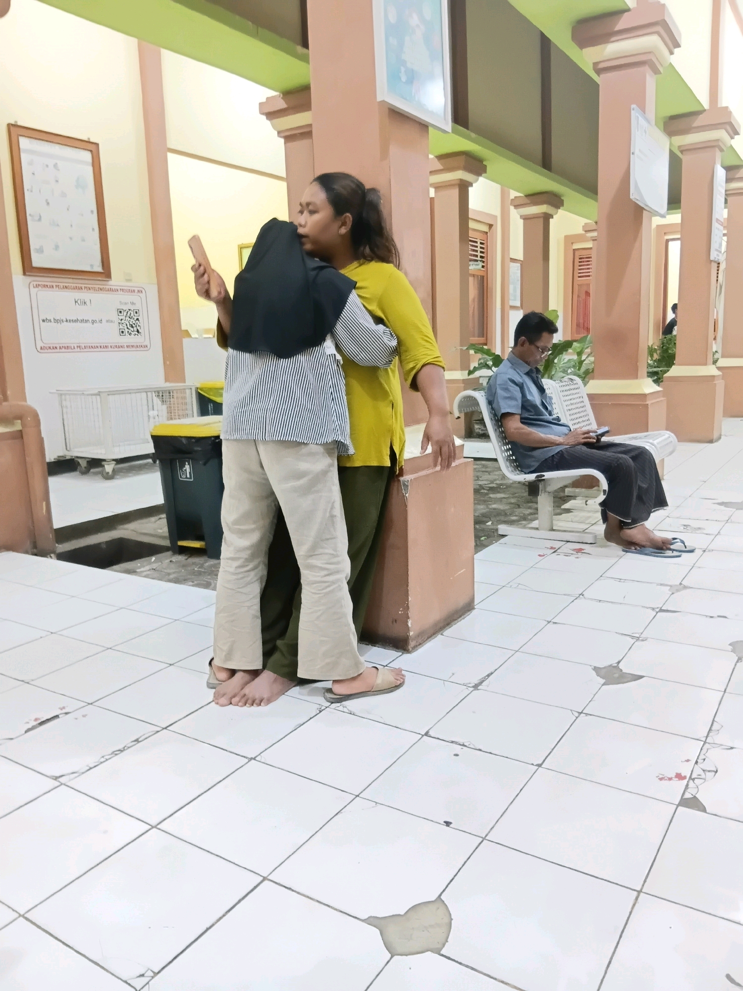 In the city of Rengel, Indonesia, an image depicts two individuals in close embrace, standing on a tiled floor in what appears to be a waiting area. One person, wearing a yellow top and olive green pants, holds a phone. The other, dressed in a black hijab and a striped shirt with beige pants, is being embraced. Their feet are bare or clad in sandals. In the background, a man wearing glasses and a blue shirt sits on a white bench, looking at his phone. The environment features beige pillars and walls, with a green ceiling. A trash can and a wheeled cart are visible, as well as a framed poster on the wall. The overall scene suggests a public space, possibly a clinic or administrative building.