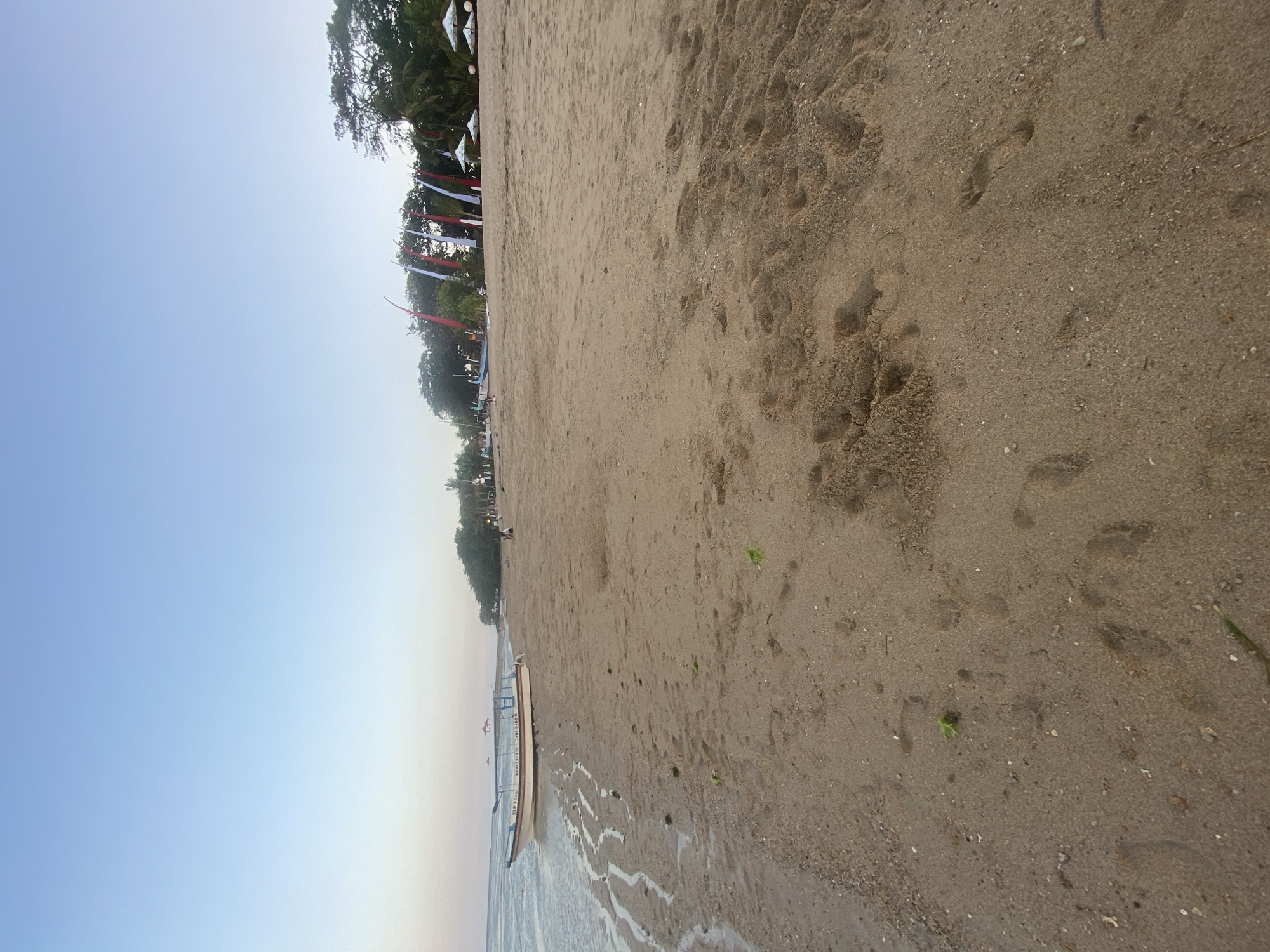 This is an eye-level, medium shot of a sandy beach in Denpasar, Indonesia, at the water's edge during daylight. The sand is wet and shows footprints. In the foreground, the shallow water laps at the shore, leaving a thin film of water on the sand. A long, narrow white boat with blue trim is beached on the wet sand to the left of the frame. In the background, a line of trees and vegetation marks the edge of the beach. Several white and red flags are planted along the treeline, with two white umbrellas visible behind them. A few people are scattered along the beach in the distance, and some appear to be near the treeline, possibly sitting or walking. The sky is a clear, pale blue, transitioning to a lighter shade near the horizon. The overall atmosphere is tranquil and serene.