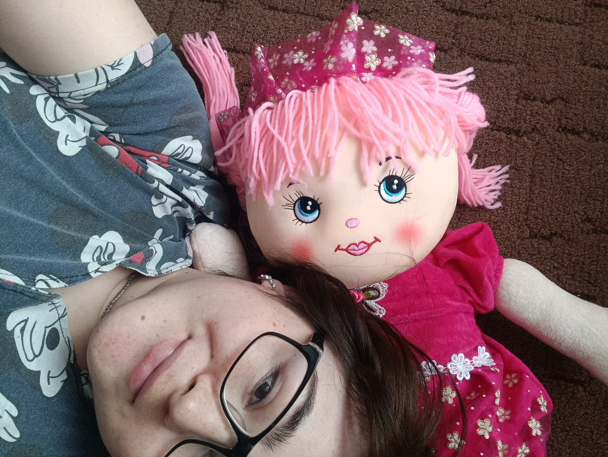 A woman with glasses is lying on a brown surface next to a doll. The woman has dark hair and is wearing a blue and gray shirt with Mickey Mouse images. She is also wearing a silver chain. The doll has pink yarn hair and a pink dress with white and gold flowers. It has a matching pink crown. The background is a textured brown surface, likely a carpet or rug. The location is not identifiable as Jelpachiha, Russia from this image.