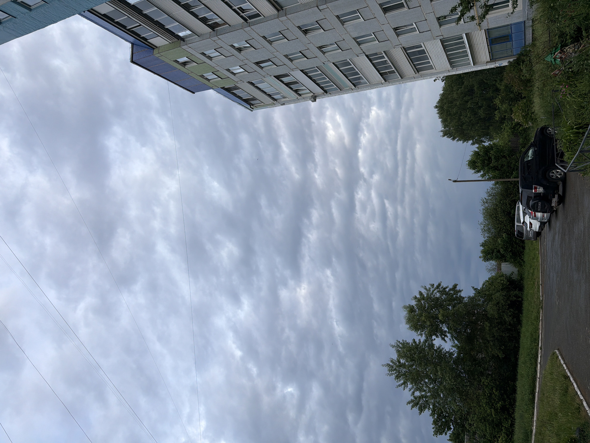 The image shows a view looking upwards towards a cloudy sky in what appears to be a residential area. The corner of a multi-story building is visible at the top of the frame, with rows of windows suggesting it is an apartment complex. The building's facade has a gray and blue color scheme. Along the right side of the image, there is greenery, possibly trees and bushes, bordering a paved area. Several cars are parked along the edge of this pavement. Power lines are visible stretching across the sky. Given the location tag, this scene is likely in or near the city of Chaikovsky, Russia. There are no people immediately visible in the image.