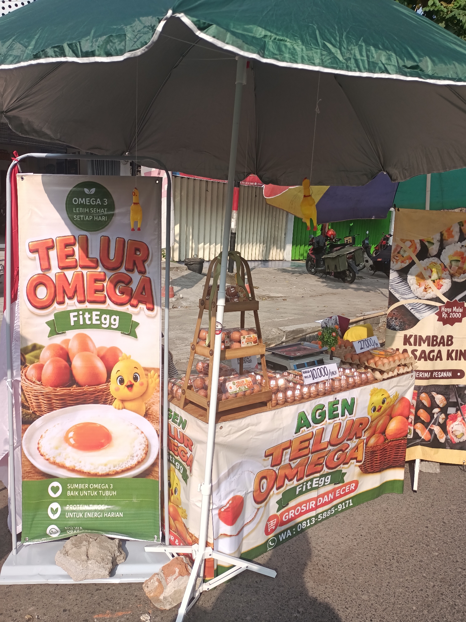 A street food stall is set up outdoors, under a large green umbrella. The stall is selling eggs and kimbap, with prominent banners advertising "TELUR OMEGA FitEgg" (Omega Eggs FitEgg) and "KIMBAB KANAGA KIN" (Kimbap Kanaga Kin). A small, yellow cartoon chicken character is featured on the "TELUR OMEGA" banner. There are several stacks of eggs in cardboard cartons and a basket of eggs on display. A scale is visible on the counter, suggesting sales. In the background, two motorcycles are parked, and there are buildings with corrugated metal siding. The ground is paved with asphalt. The scene appears to be in a market or busy street area. The presence of the Indonesian language on the banners and the type of food sold indicate the location is likely in Indonesia, specifically Pare, based on the context provided.