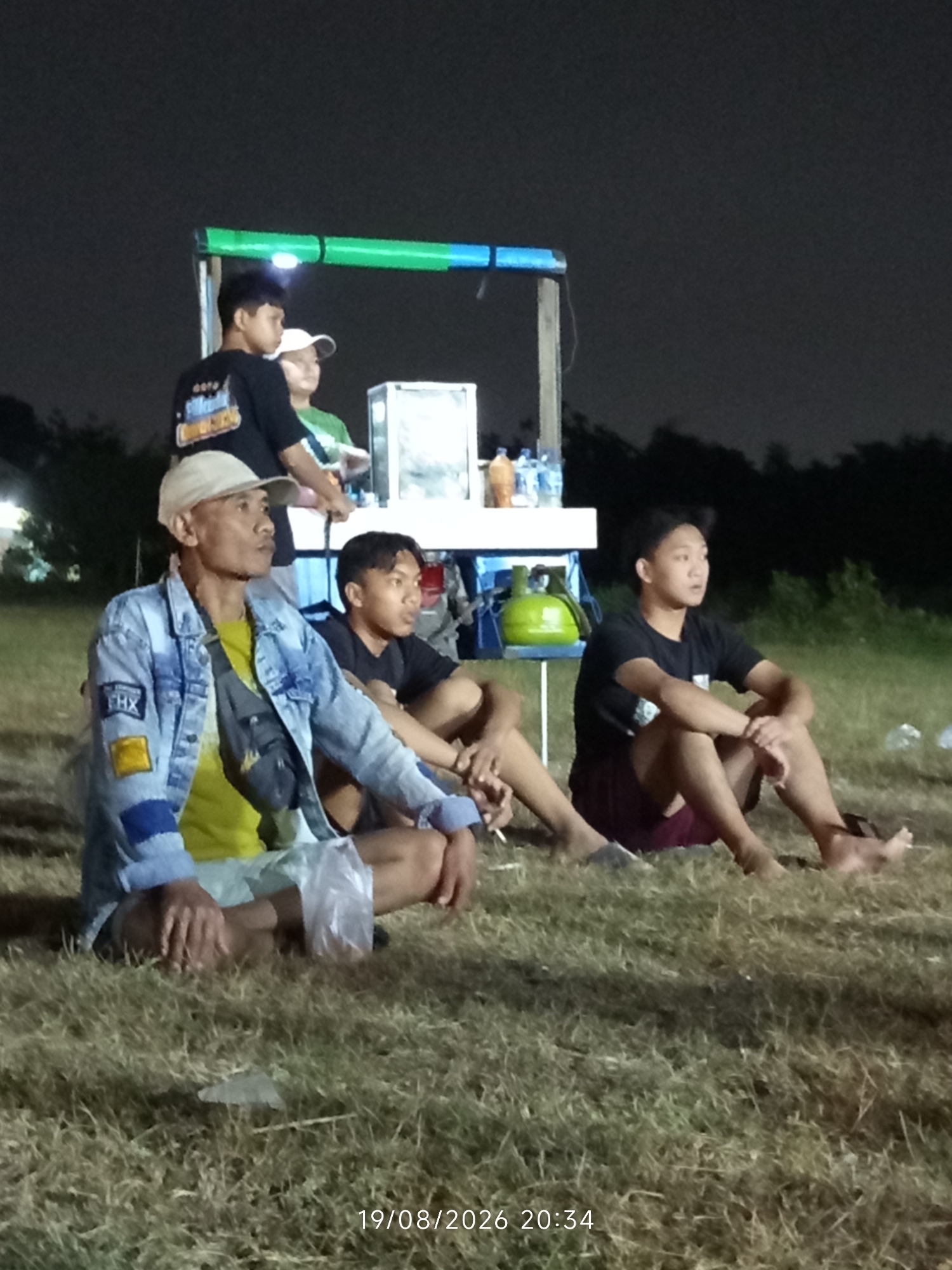 The image depicts a nighttime scene in Canggu, Indonesia, featuring a group of individuals gathered outdoors. In the foreground, a man wearing a light-colored baseball cap and a blue denim jacket over a yellow shirt sits on the grass. He has a dark-colored sling bag across his chest. To his right, two young men are seated on the ground. One is wearing a black t-shirt and burgundy shorts, and the other is wearing a black t-shirt. Behind them, two more individuals are standing near a food cart. One, wearing a black t-shirt with a graphic design, stands next to another person in a light-colored cap and a green shirt. The food cart, with a green and blue awning, is illuminated by a light. Bottles of water and a green gas canister are visible on a table attached to the cart. The ground is covered in dry grass, and the background is dark, suggesting it is nighttime. A timestamp in the lower right corner reads "19/08/2026 20:34".
