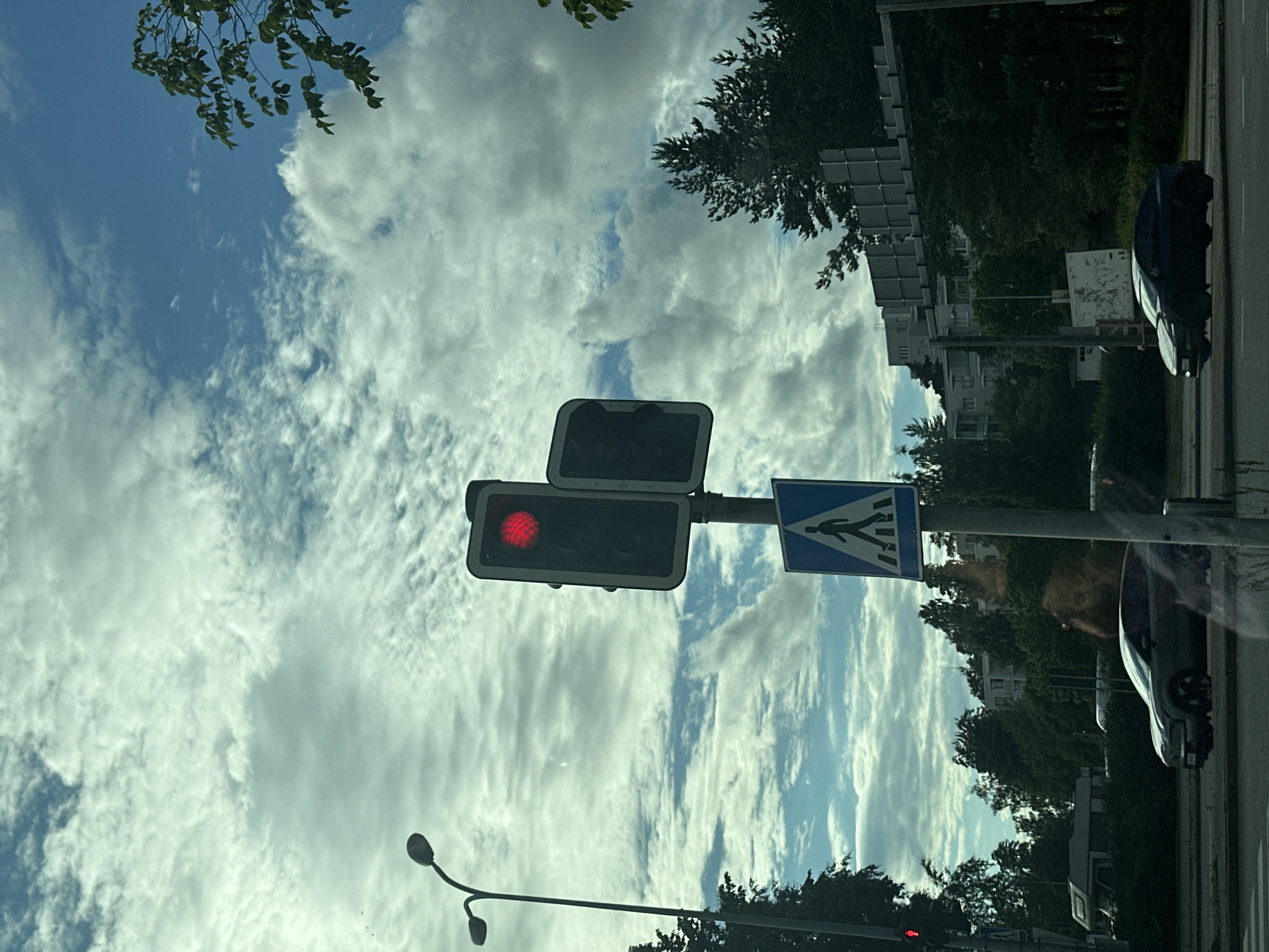 The image shows a street scene with a traffic light and a pedestrian crossing sign. The traffic light is red, indicating that vehicles should stop. The pedestrian crossing sign, a blue square with a white silhouette of a pedestrian, is visible to the right of the traffic light. In the background, there are trees, buildings, and a street. A dark-colored car is visible on the right side of the road. The sky is blue with scattered white clouds. The overall impression is that it is daytime, and the weather is clear. The image is oriented vertically, with the sky at the top and the ground at the bottom. There is no visible text in the image.