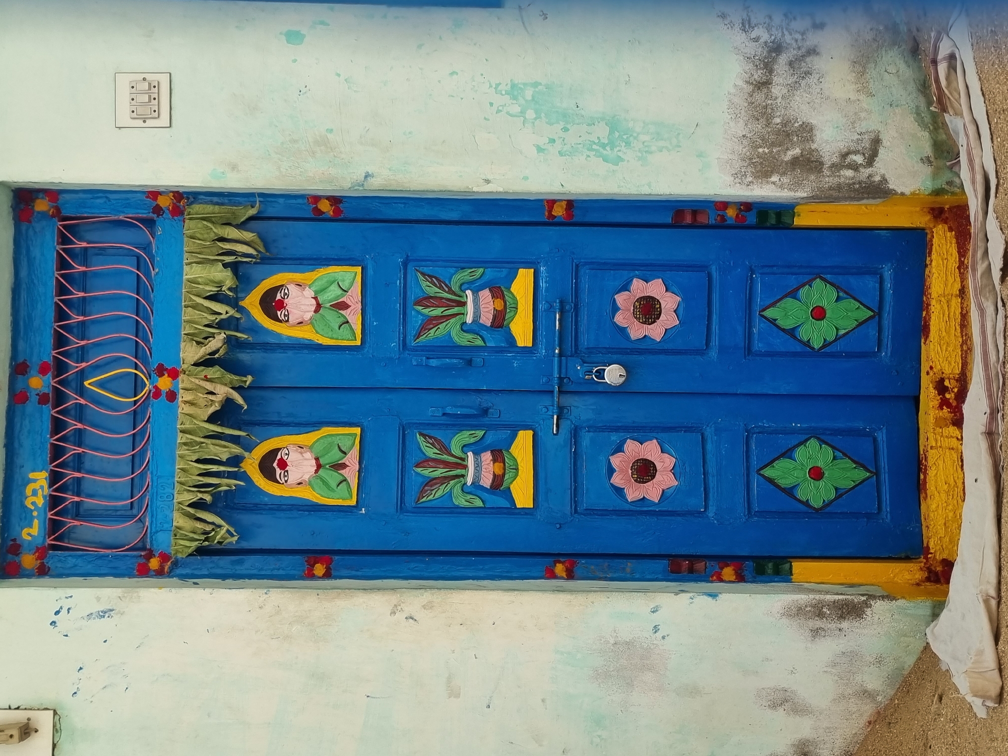 This image depicts a brightly painted blue wooden double door set into a faded light blue/green wall in Pedda Kammavaripalli, India. The door, likely an entrance to a residence, features a yellow-painted number "2.231" at the top. Below this, a pink metal grillwork with a central yellow teardrop shape is visible. A string of green leaves and small red/yellow decorative elements hangs horizontally beneath the grill.
The door panels are adorned with multiple carved or painted motifs. The upper two panels each display a stylized human figure with a yellow veil, green attire, a red bindi on the forehead, and hands pressed together in a gesture of reverence. The two middle panels each feature a light pink pot (kalash) with red stripes, containing green leaves, resting on a yellow base. The door is secured at its center by a silver padlock on a metal latch. The lower panels display stylized pink five-petal flowers, and the bottommost panels depict green rhombus shapes with red centers, representing flowers. The door's frame and threshold are painted yellow, with some red staining near the bottom.
The surrounding wall is light blue/green, showing signs of wear, discoloration, and dirt accumulation, particularly at the base. To the right of the door, an off-white electrical switchboard with three white switches is mounted on the wall. On the ground to the left of the door, a pile of light brown sand or dirt is visible. Directly in front of the door's threshold lies a crumpled piece of white fabric, possibly a cloth, with red stripes. No individuals are present in the scene, and no specific actions are occurring.