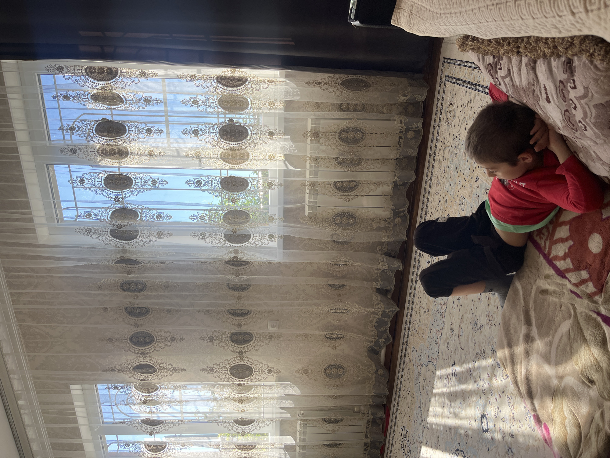 A young child is lying on a bed in a room. The child is wearing a red shirt and black pants. The bed is covered with a patterned blanket. A window with ornate curtains is visible in the background. The location is Kaspiysk, Russia.