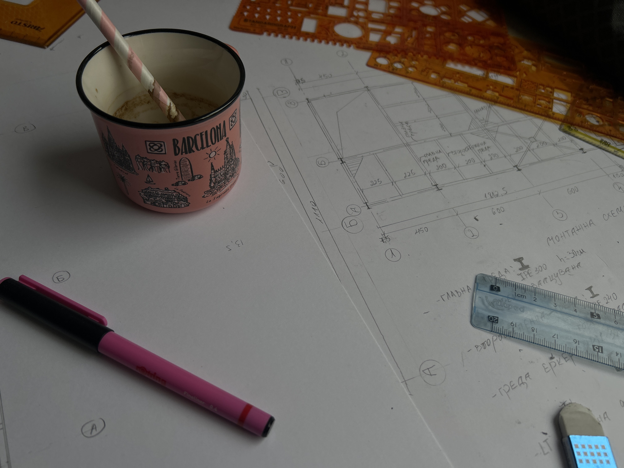 The image presents an organized, but cluttered workspace with various architectural design tools and materials. The scene includes architectural plans and blueprints scattered across a white surface, along with a yellow architectural scale at the top of the image. There is a pink "Barcelona" mug with a striped straw in it, a pink marker pen, a ruler, and a blue eraser. The plans appear to be hand-drawn and feature architectural notations and measurements in what appears to be Bulgarian. The focus is on the intersection of design work and daily routines, captured through the juxtaposition of technical tools with a personal coffee mug. The setting implies an architectural design studio or a student's workspace.