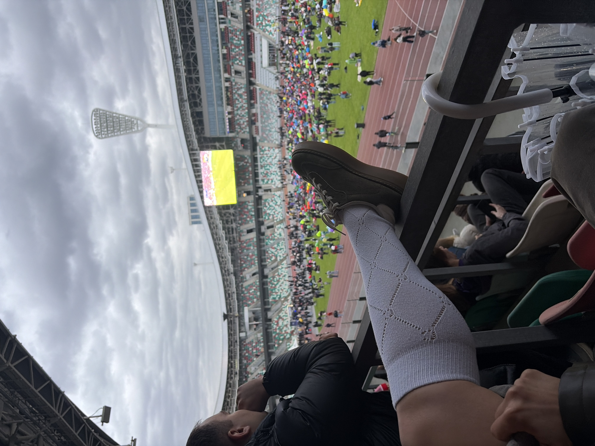 The image was taken in Minsk, Belarus, at a stadium filled with spectators. The photograph is taken from a high vantage point, likely within the stands. A man's leg, clad in a white patterned sock and a brown shoe, is propped up on the railing. A large video screen and a distinctive metal structure, possibly a floodlight support, are visible in the background. The sky is overcast, suggesting a cloudy day. The stands of the stadium are densely packed with people. There's an umbrella in the foreground on the right side.