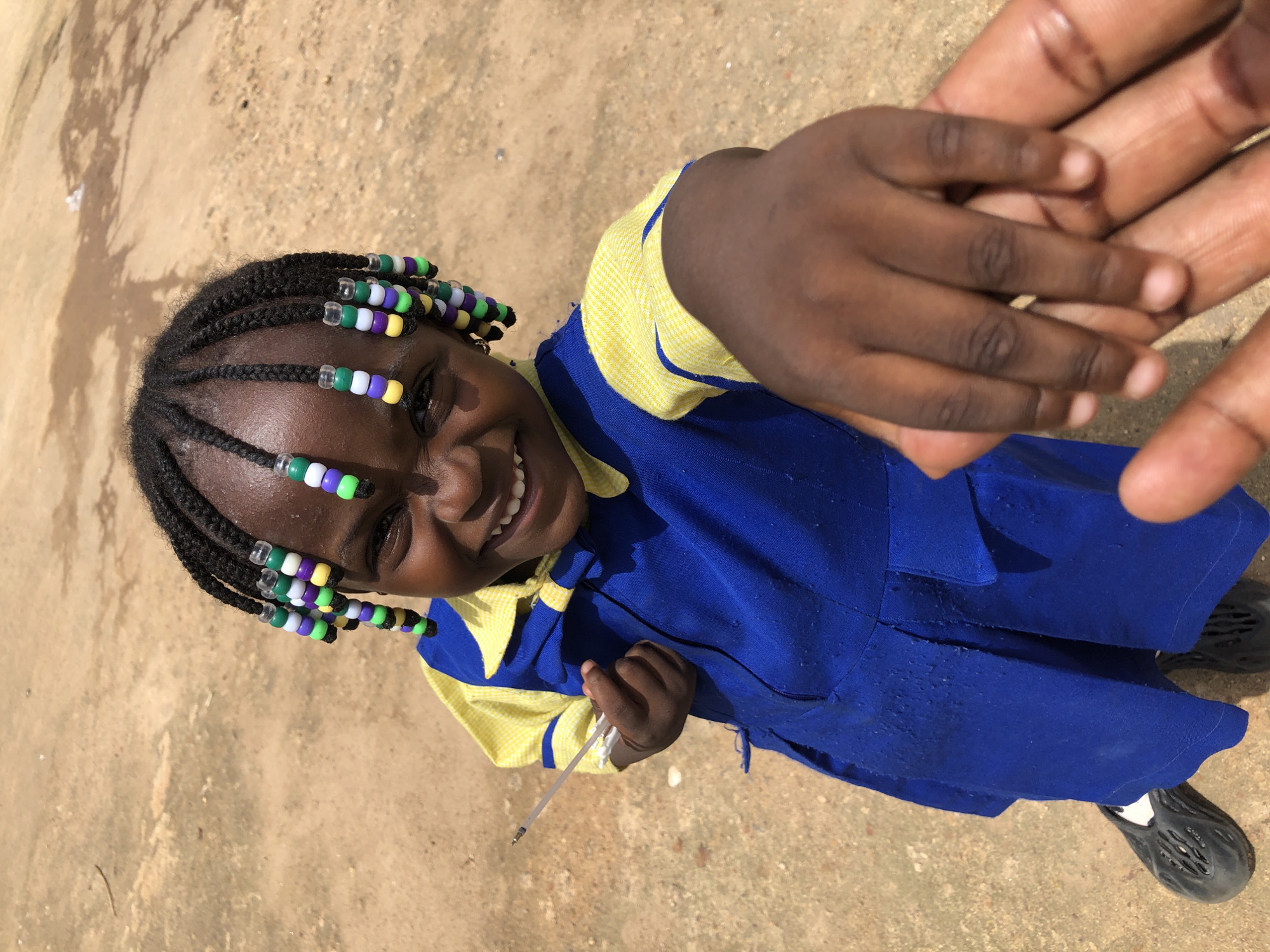 A close-up, high-angle outdoor shot shows a young Black girl with braided hair decorated with colorful beads, wearing a blue school uniform with a yellow checkered collar and sleeves. She is smiling broadly and extending her right hand toward the camera, holding a pen. The ground around her is a dusty, dry earth, with shadows cast from an unseen object. Her left hand is visible in the lower left of the frame, and another hand is holding hers in the lower right. The image appears to have been taken in Jos, Nigeria, given the common school uniform style and the dry, dusty environment indicative of the region.