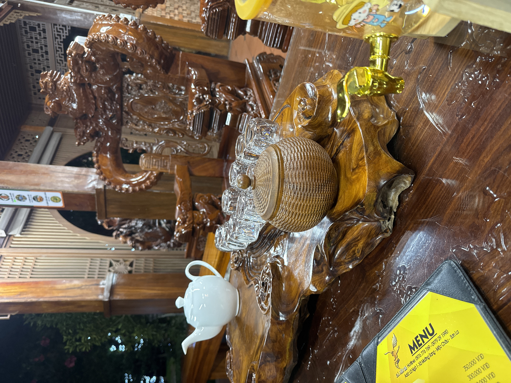 In Mộc Châu, Vietnam, a wooden table displays a tea set consisting of a white teapot, several clear glass teacups arranged in a row, and a small, woven lidded container. A decorative wooden tray, carved with organic, flowing lines, supports the tea set. In the background, an ornate, carved wooden chair with dragon motifs suggests an interior space. To the right, a portion of a clear dispenser with a gold spigot is visible, featuring a cartoon character on its side. A yellow menu with black text is partially visible in the foreground, resting on the table. The table surface is dark, polished wood, with some scattered water droplets. The lighting is dim, suggesting an evening setting, with some ambient light visible through a wooden-latticed background structure.