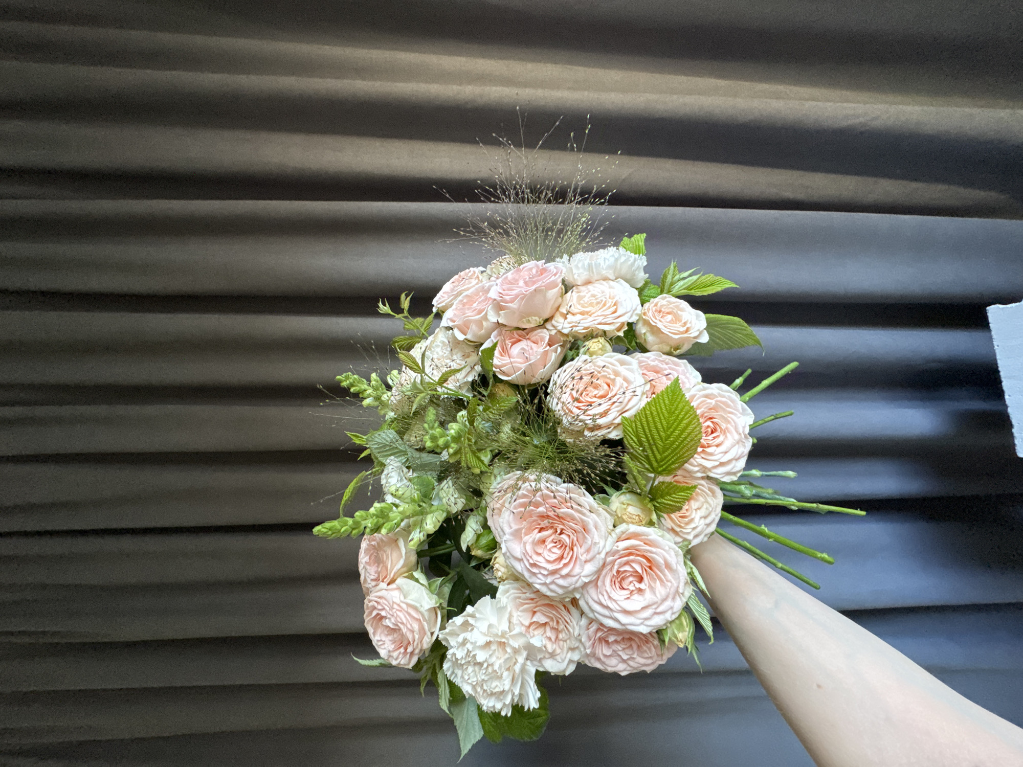 A bouquet of pink roses and white carnations is held by a person's arm, in front of a dark gray curtain. The bouquet is lush and full, with various green foliage and delicate wispy grasses. The roses are a soft peachy-pink hue, with tightly packed petals. The carnations are a creamy white with ruffled edges. The curtain behind the bouquet is draped in vertical folds, creating a textured background. The lighting is soft and diffused. There is no visible indication of the specific location within Izhevsk, Russia, in this image.