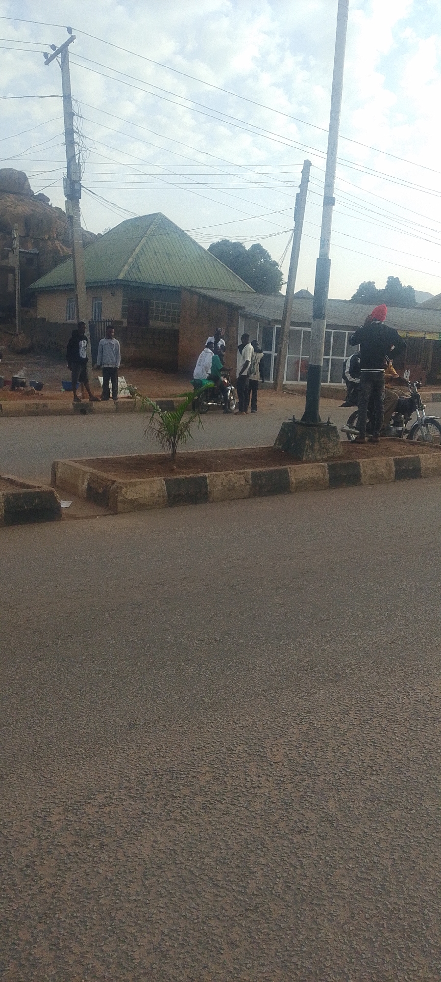 The image depicts a street scene in Gura Top, Nigeria. A group of individuals are gathered near a building with a green, pitched roof. Several people are standing around, some are on motorcycles. The road in the foreground is paved and has a concrete curb with black and white markings. Power lines and poles are visible overhead, indicating a utility infrastructure. The background shows a rocky outcrop and trees, suggesting a semi-urban or rural setting. The sky is cloudy.