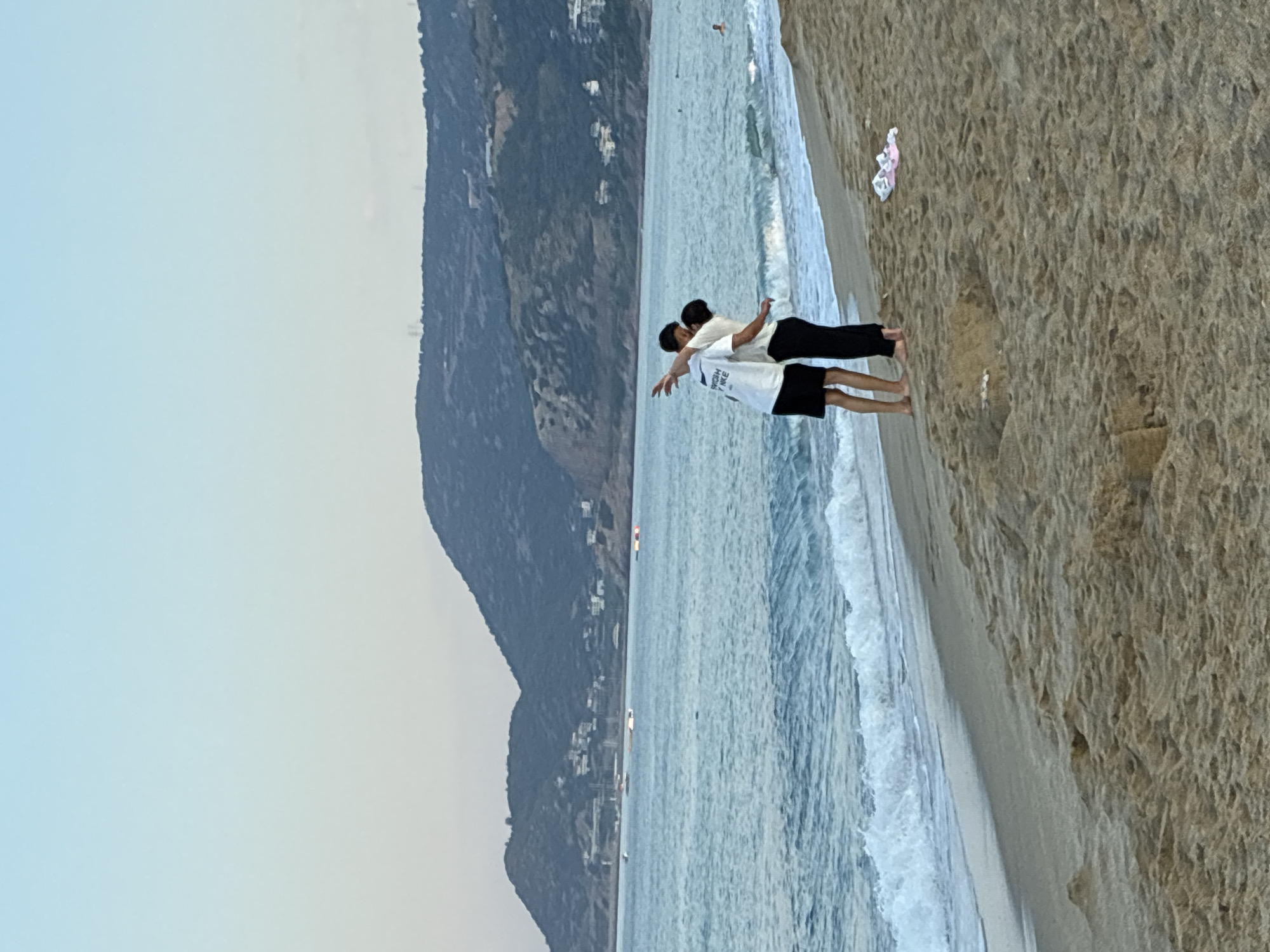 Two individuals are standing on a sandy beach in Alanya, Turkey, facing the sea. The person on the left wears a white t-shirt with text and black shorts, while the person on the right wears a white t-shirt and black pants. They appear to be embracing or interacting closely. The sea is visible in the background with gentle waves and a few boats on the horizon. Hills with scattered buildings and vegetation form the backdrop, characteristic of the Alanya coastline. The sky is a soft gradient of blue and pink, suggesting either sunrise or sunset. A pair of pink flip-flops and other small objects are on the sand near the individuals.