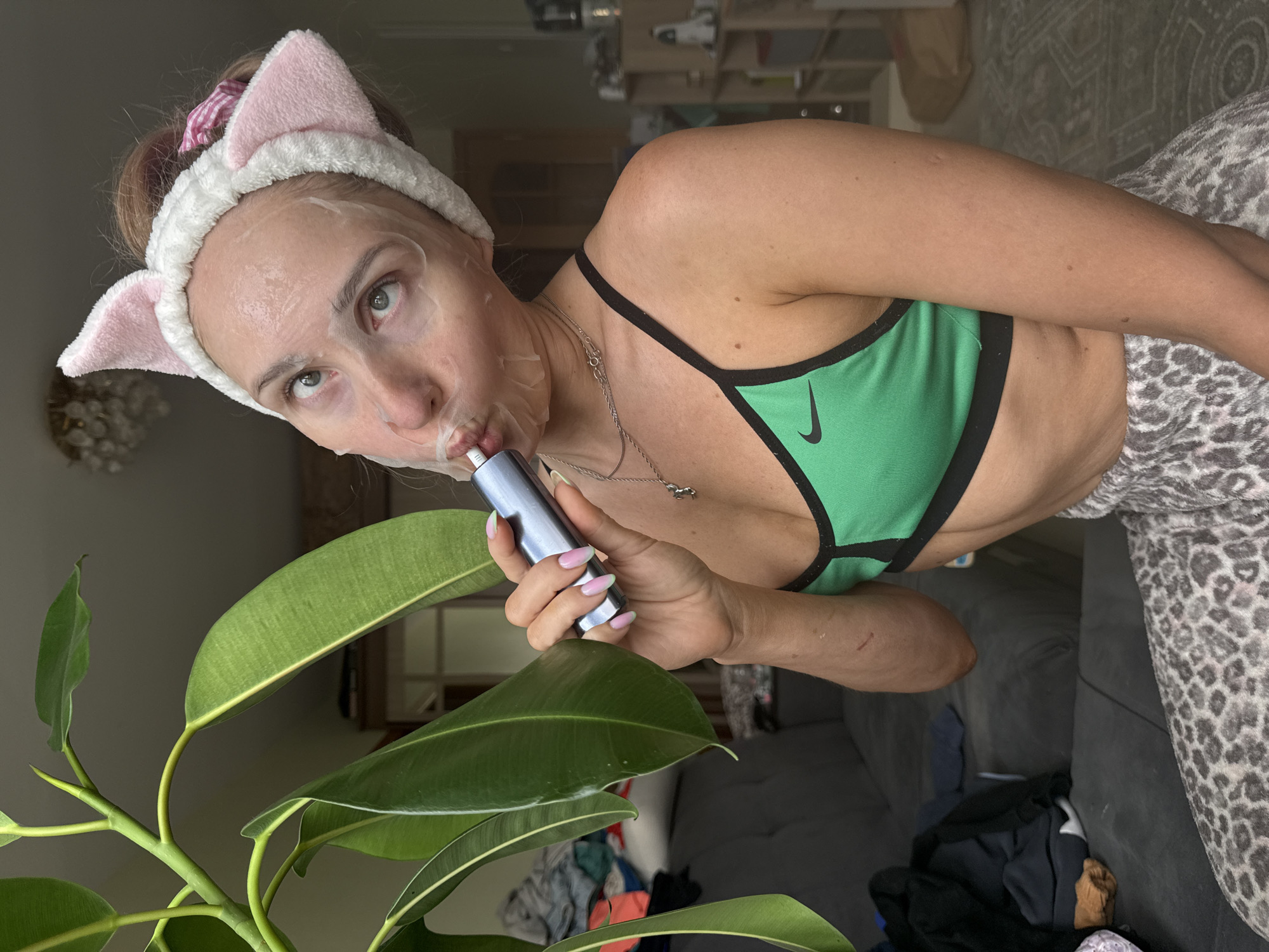A young woman with a sheet mask on her face and a pink cat-ear headband is holding a silver cylindrical device to her mouth. She is wearing a green sports bra and leopard-print pajama pants. The woman is in an indoor setting, likely a living room, with a couch and a plant in the background. The background suggests a domestic environment with some clutter. The image does not contain enough information to determine the location in Chaikovsky, Russia.