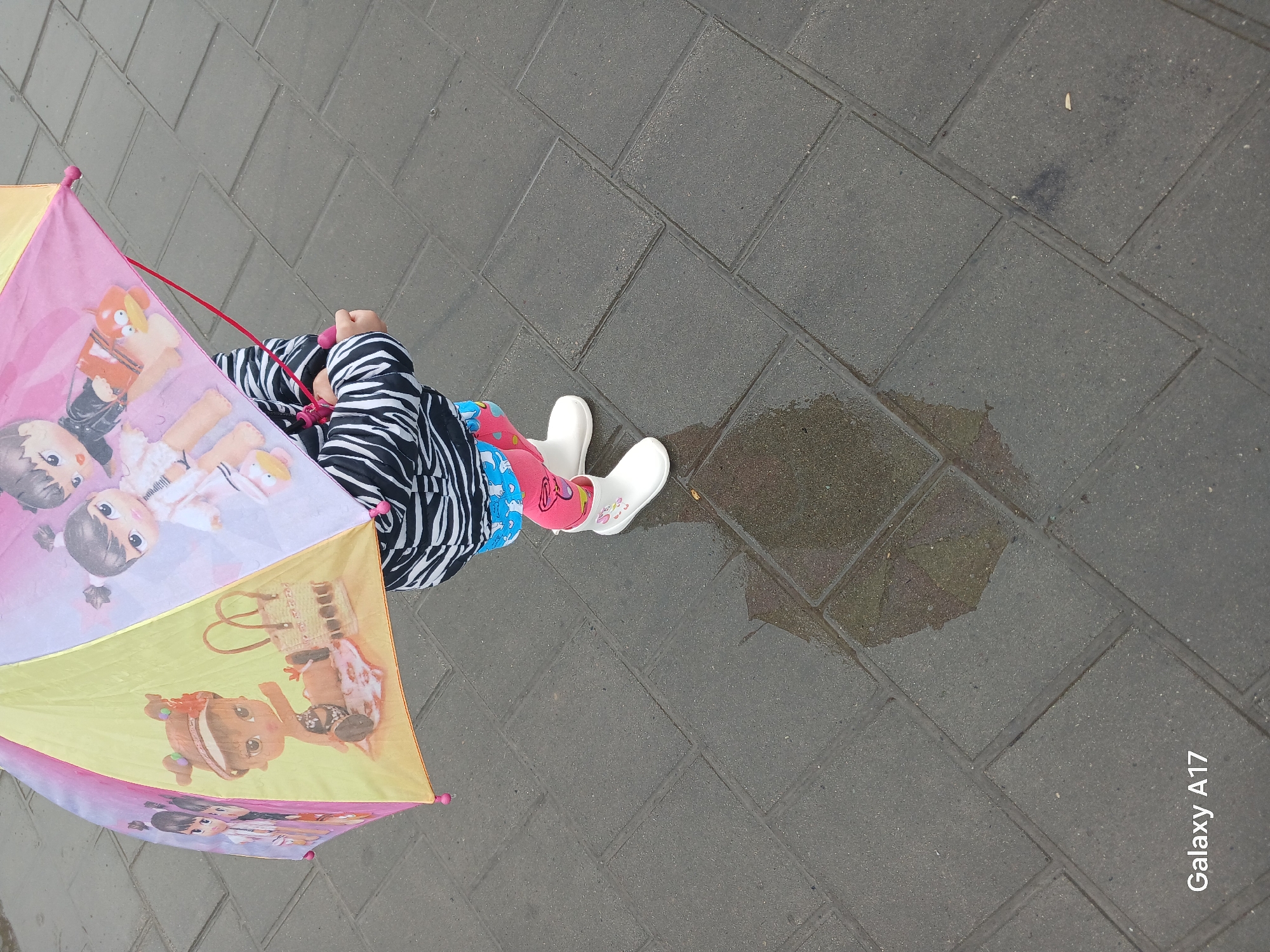 A child, facing away from the camera, stands on a gray paved surface under a cartoon-printed umbrella. The child wears a black and white striped jacket, pink and blue patterned leggings, and white boots. A puddle reflects an image of the umbrella on the pavement. The phrase "Galaxy A17" appears in the lower right corner. The image was taken outdoors, potentially in Yekaterinburg, Russia, as the location was specified in the prompt.