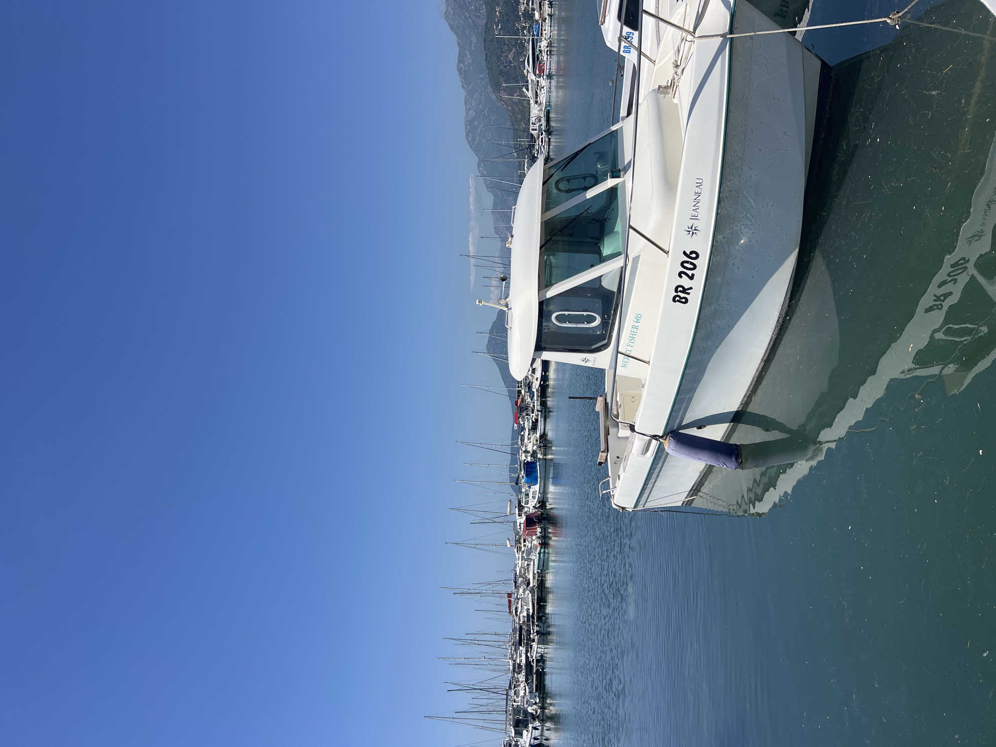 The image depicts a marina in Bar, Montenegro, with numerous boats moored in the water. In the foreground is a white motorboat labeled "BR 206 JEANNEAU MERRY FISHER 605." Its hull is reflected in the dark water. The background shows rows of sailboats and motorboats, with masts extending upwards. Mountains are visible in the distance under a clear blue sky. The scene is captured in daylight, indicating sunny weather.