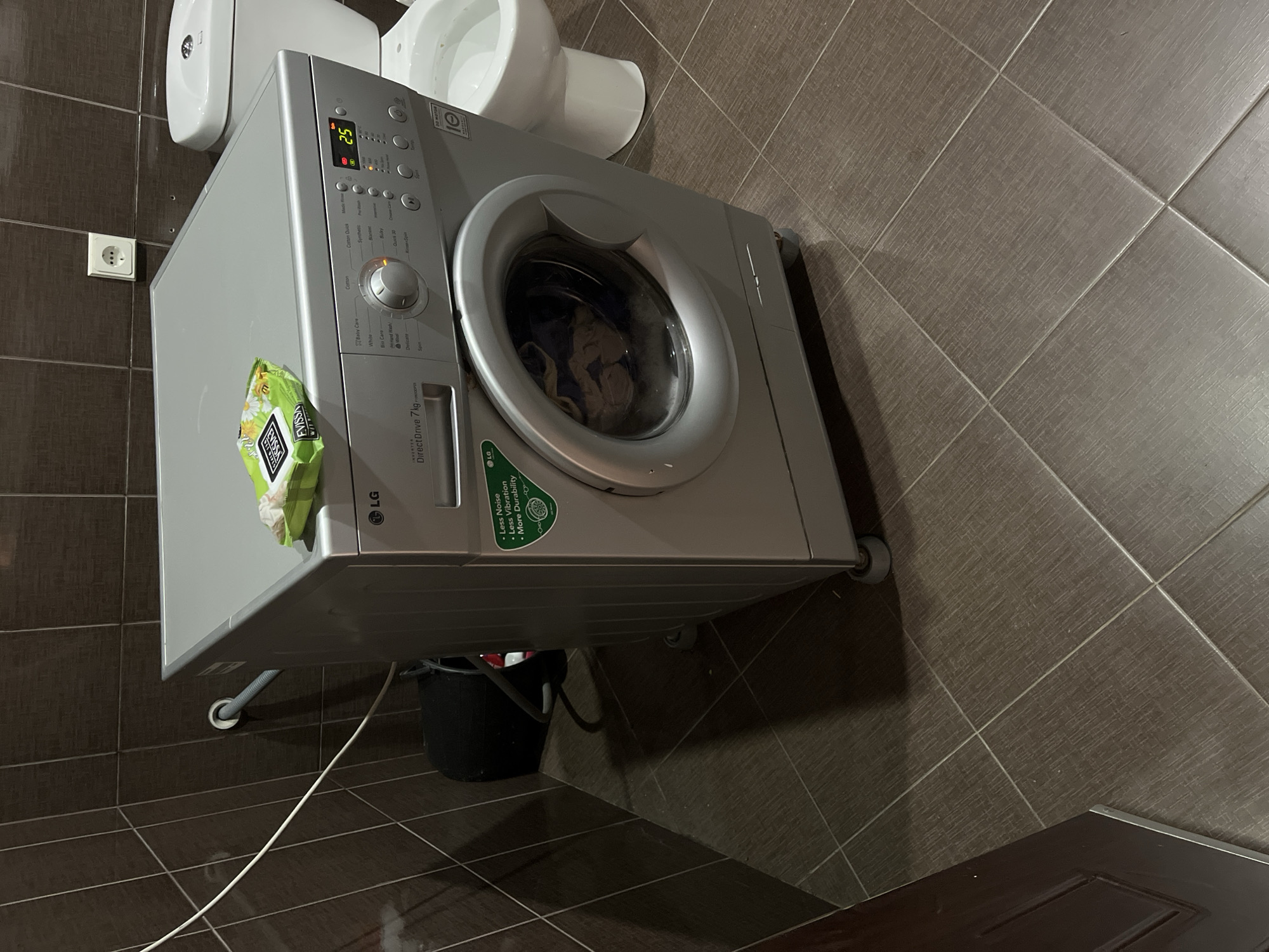 An LG DirectDrive 7kg front-loading washing machine is positioned in a tiled bathroom. On top of the washing machine is a package of wet wipes. To the right of the washing machine is a white toilet. The walls are tiled with dark brown tiles. A white electrical outlet is visible on the wall above the washing machine. A dark object, possibly a bucket, is located behind the washing machine on the left side of the frame. A white power cord is visible on the wall and floor. A closed dark brown door is visible in the lower left corner of the image.