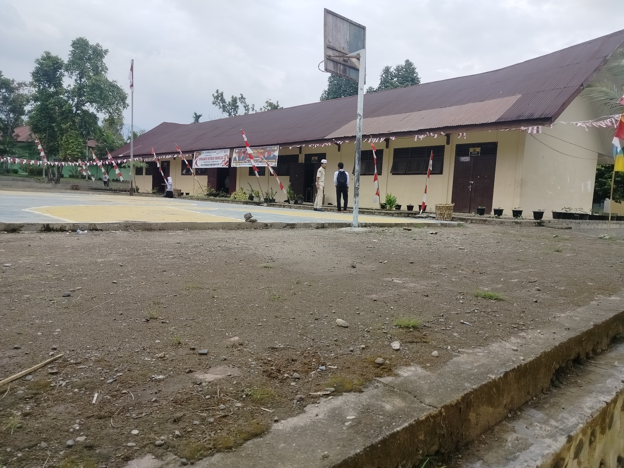 The image shows a side view of a single-story building with a brown corrugated metal roof. The building is painted a light yellow and has several windows and two large double doors. Indonesian flags are draped along the front of the building, and small red and white pennant banners are strung across the facade. Two individuals, one in a light-colored uniform and the other in a dark shirt and pants, stand near the entrance. In the foreground, there is a dirt and gravel area with some scattered stones and sparse vegetation. A concrete curb runs along the bottom edge of the image. A basketball hoop with a weathered backboard is visible to the right of the building. The background shows trees and a cloudy sky. The scene appears to be a schoolyard in Lawebekung, Indonesia, likely decorated for a national holiday.