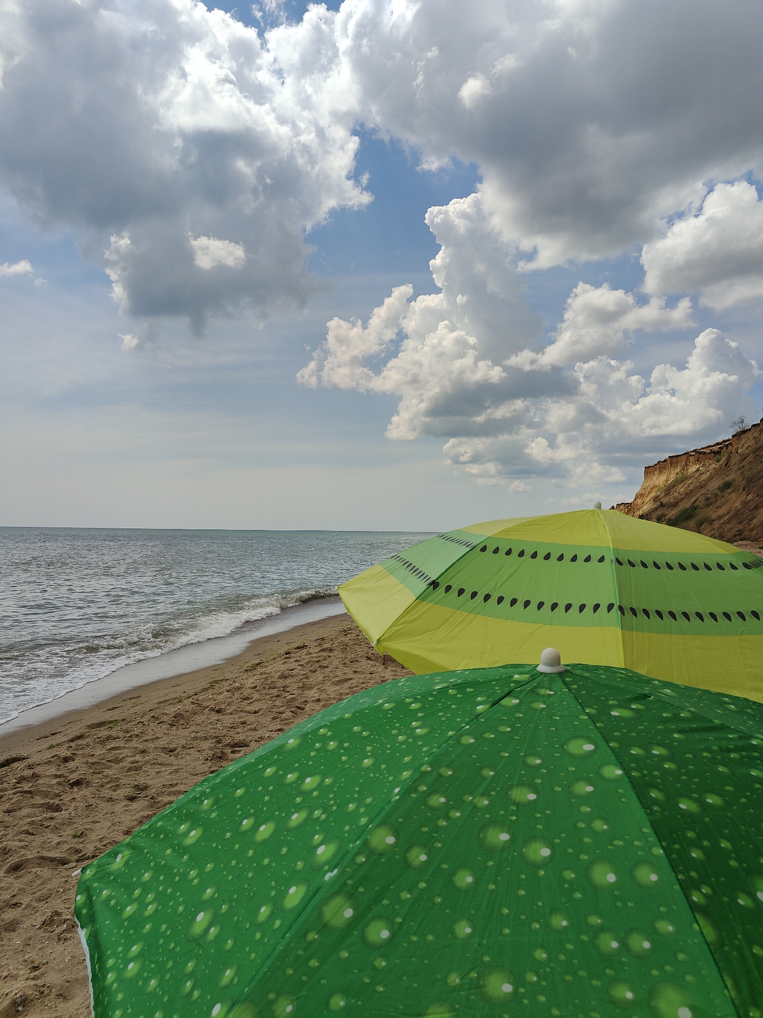 Two colorful beach umbrellas are set up on a sandy beach next to the sea. The umbrella in the foreground is green with a pattern of white circles resembling water droplets. Behind it, a yellow umbrella with horizontal green stripes and black teardrop shapes is visible. The sea stretches out to the horizon under a partly cloudy sky. In the background, a steep cliff face rises from the beach. The scene appears to be located in Sanzhiika, Ukraine, a coastal village known for its beaches and cliffs. No people are present in the frame.