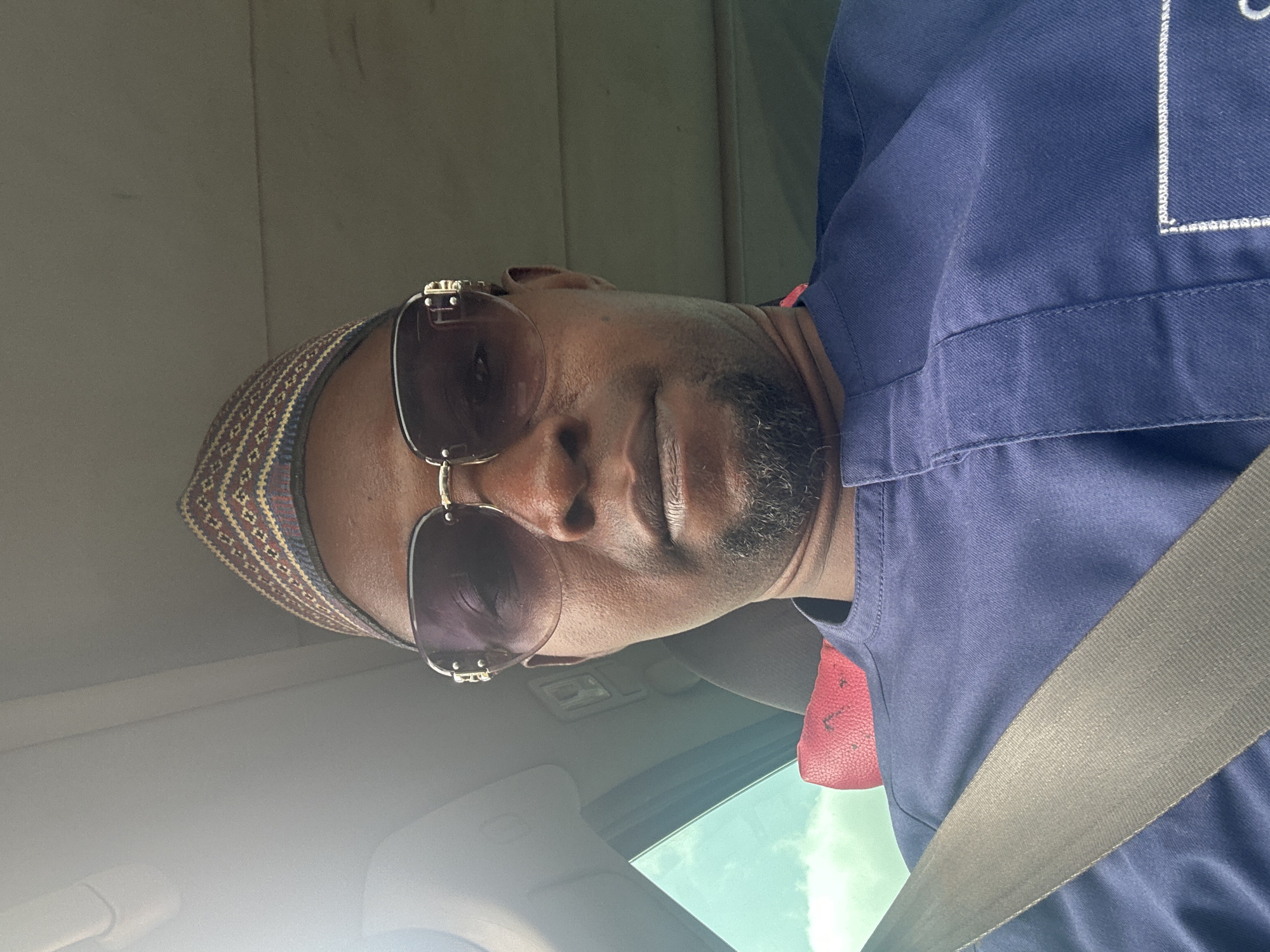 A man is wearing sunglasses and a patterned kufi cap while sitting in a car, secured with a seatbelt. He's wearing a blue shirt with some white detailing on the shoulder. The image seems to be taken from inside the car, looking up at the man. The location appears to be a car in the city of Unknown, Unknown.