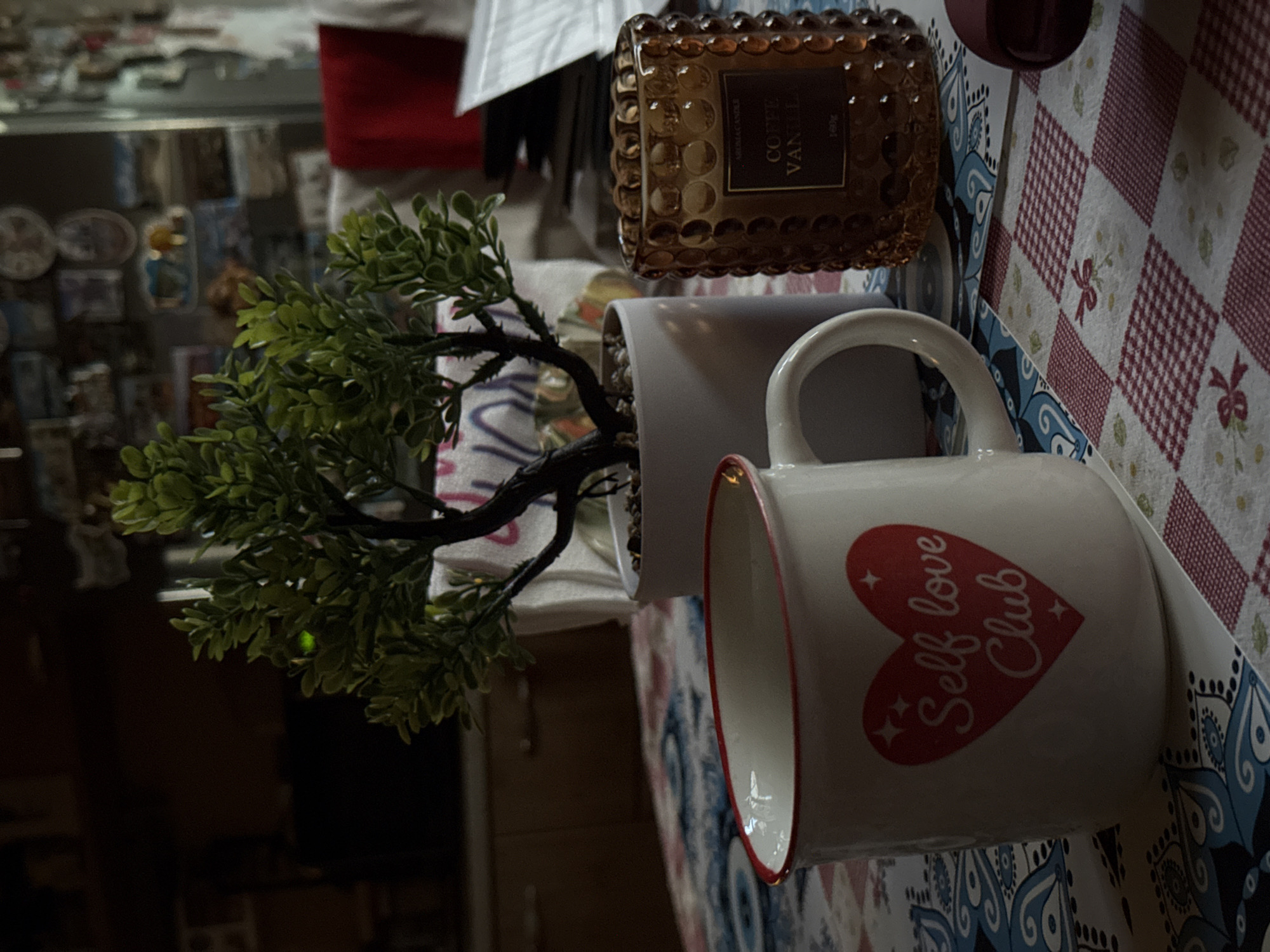 The image depicts a close-up of a table setting. In the foreground, a white ceramic mug with a red rim is prominently featured. The mug has a red heart design on its side, with the text "Self love Club" written inside. To the right of the mug is a textured amber glass candle holder with a label that reads "COFFEE VANILLA." Behind the mug, a small artificial potted plant with green leaves is visible. The table is covered with a patterned tablecloth, which appears to have a blue and white floral design in the foreground and a red and white checkered pattern further back. The background is dark and out of focus, showing glimpses of what appear to be kitchen cabinets and shelves with various items. There is no indication of the city of Pleven, Bulgaria, within the image itself.