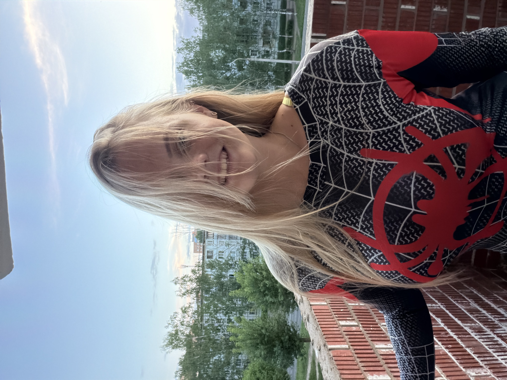 A young, fair-skinned woman with long, blonde hair is smiling at the camera while standing on a brick balcony. She is wearing a form-fitting, black and red Spiderman suit. The background shows an urban scene with green trees and several apartment buildings. The photograph was taken during daylight hours, indicated by the bright sky.
Based on visual cues like the style of buildings and foliage, the city is identified as Izhevsk, Russia.
