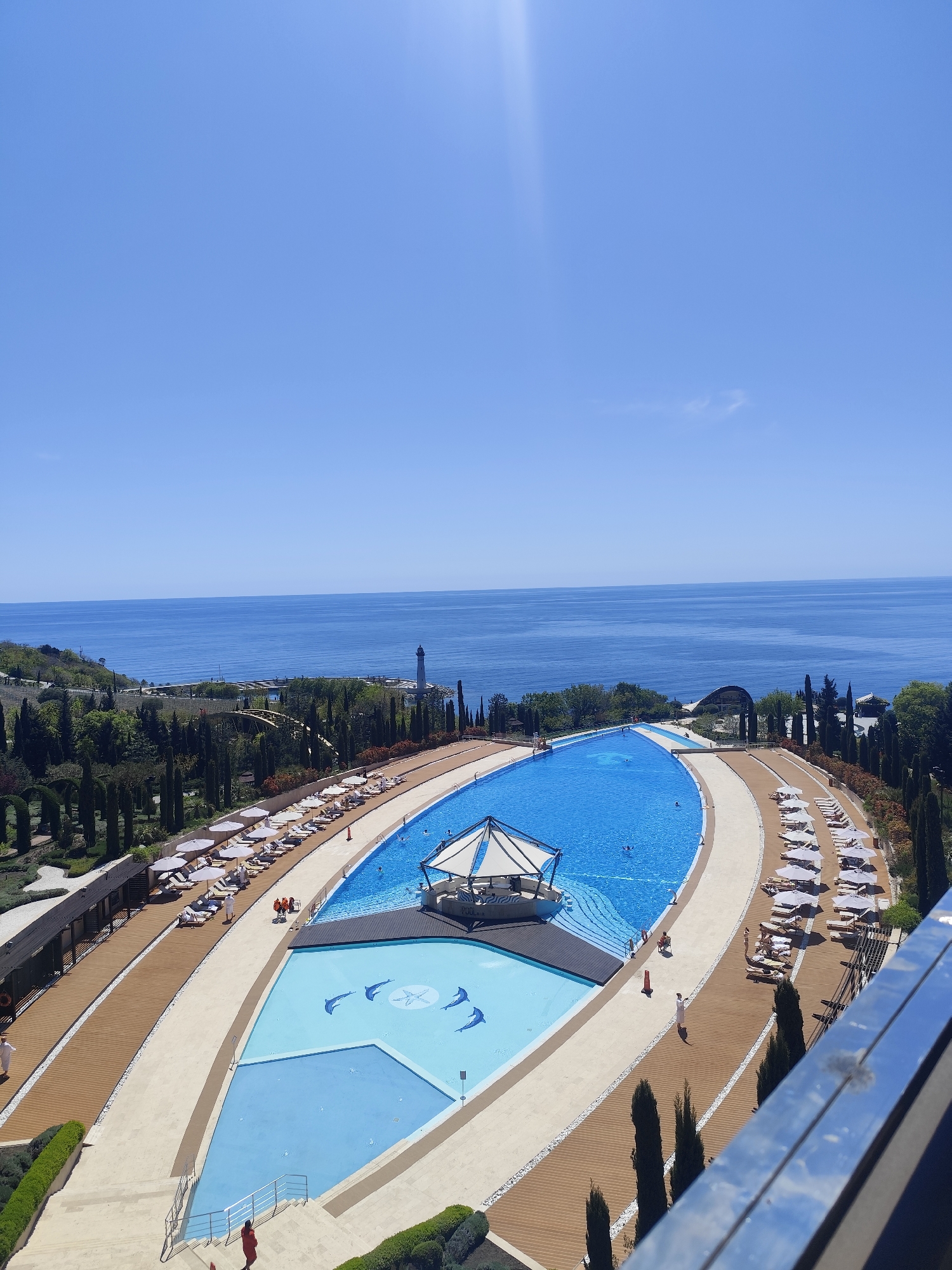 This elevated, panoramic shot captures a luxurious resort-style swimming pool complex overlooking a vast, calm blue sea under a clear, bright sky. The time of day appears to be midday, indicated by the strong sunlight and a visible lens flare in the upper left.
The main subject is a large, irregularly shaped swimming pool filled with vibrant blue water. It features a distinctive circular, white, tent-like structure, possibly a pool bar or shaded lounge, positioned within the water. Adjacent to this main pool is a shallower section of lighter blue water, adorned with outlines of a starfish in the center and several dolphins swimming around it, suggesting a decorative or children's area.
Surrounding the pools are wide pathways made of light-colored paving and darker wooden decking. These areas are extensively furnished with numerous white lounge chairs and sun umbrellas, providing ample space for relaxation. Several people are visible enjoying the facilities; some are swimming or wading in the main pool, while others are relaxing on the lounge chairs. A few individuals are seen walking on the pathways, including one person in a white garment on the wooden deck and another in red near a set of stairs.
The resort complex is built into a lush green hillside, densely landscaped with various trees, notably many tall, slender cypress-like trees that line the pathways and surrounding areas. In the distance, the deep blue sea extends to the horizon. A prominent white lighthouse or tall structure is visible on the coastline to the left, adding a nautical touch to the scenic view. The overall impression is one of leisure and tranquility at a high-end coastal destination. No discernible text is visible in the image.