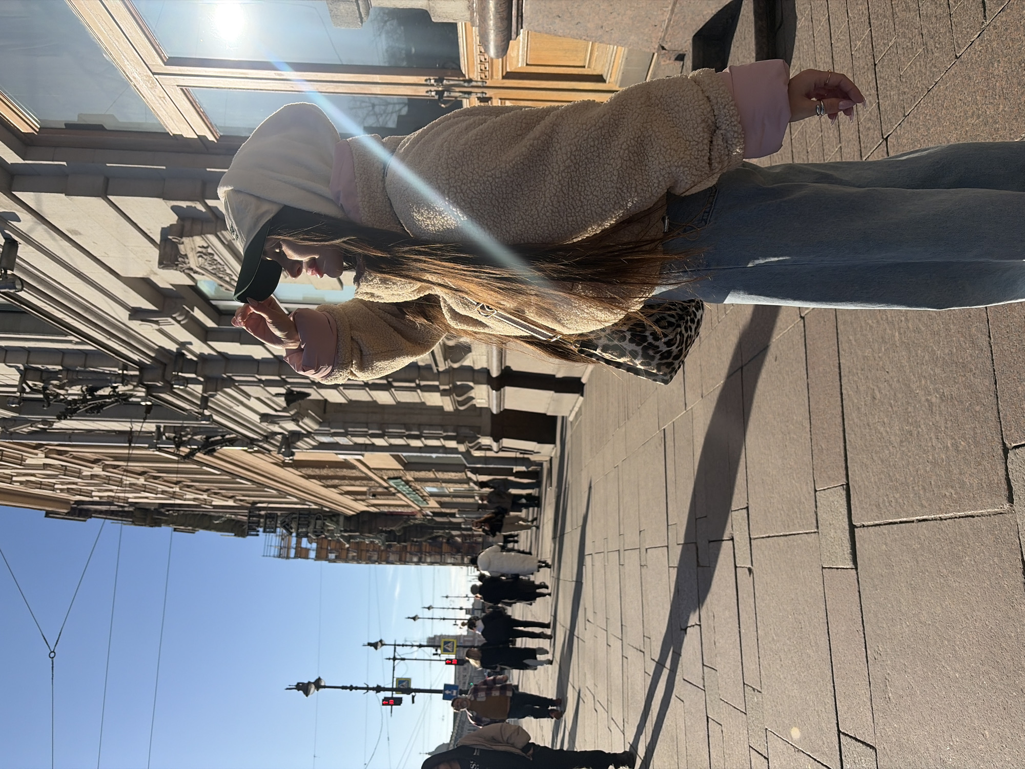 A woman with long brown hair, wearing a beige sherpa jacket with a light pink lining, blue jeans, and a baseball cap under a hoodie, stands on a paved sidewalk in Saint Petersburg, Russia. She is holding something near her face, possibly a phone or an object she is examining. A leopard-print bag hangs at her side. The background features a row of classical buildings with intricate facades, reflecting sunlight in the windows. A group of pedestrians walks in the distance along the sidewalk. Streetlights with ornate detailing line the street. The sky is clear and blue. The image is oriented at an angle, making the scene appear tilted.