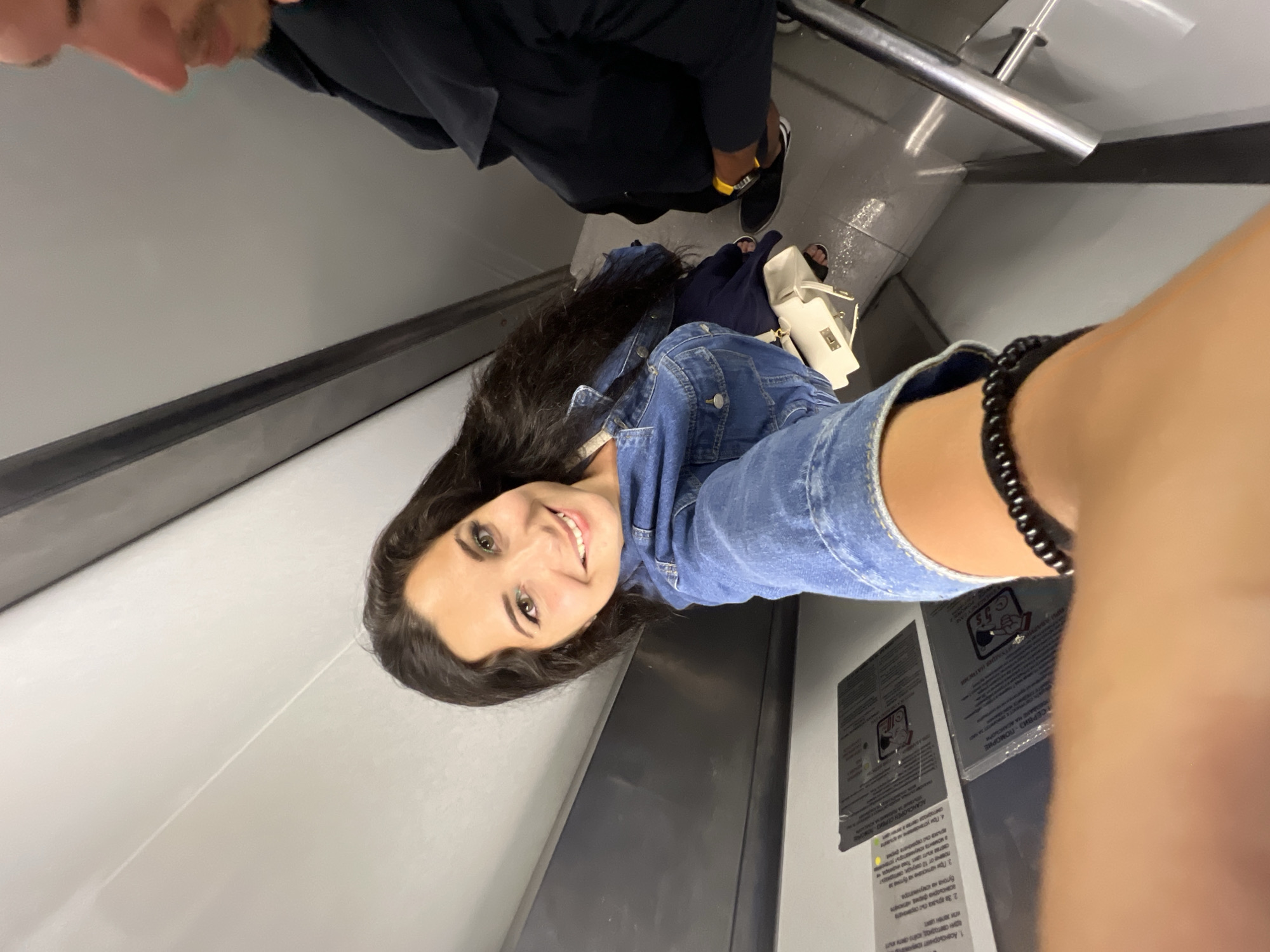 The image shows a woman taking a selfie in what appears to be a public transport vehicle, likely a bus or train. She is wearing a denim jacket and has long, dark, wavy hair. A white purse is visible on her lap, and she is wearing a black beaded bracelet on her wrist. To her right, partially visible, is a man wearing a dark jacket and a yellow watch. The interior of the vehicle is grey and utilitarian, with signs affixed to the wall. The scene suggests a moment captured during travel. The city of Pomorie, Bulgaria, is a coastal town known for its salt lakes and beaches, but this image provides no specific visual indicators to confirm its location within the city.