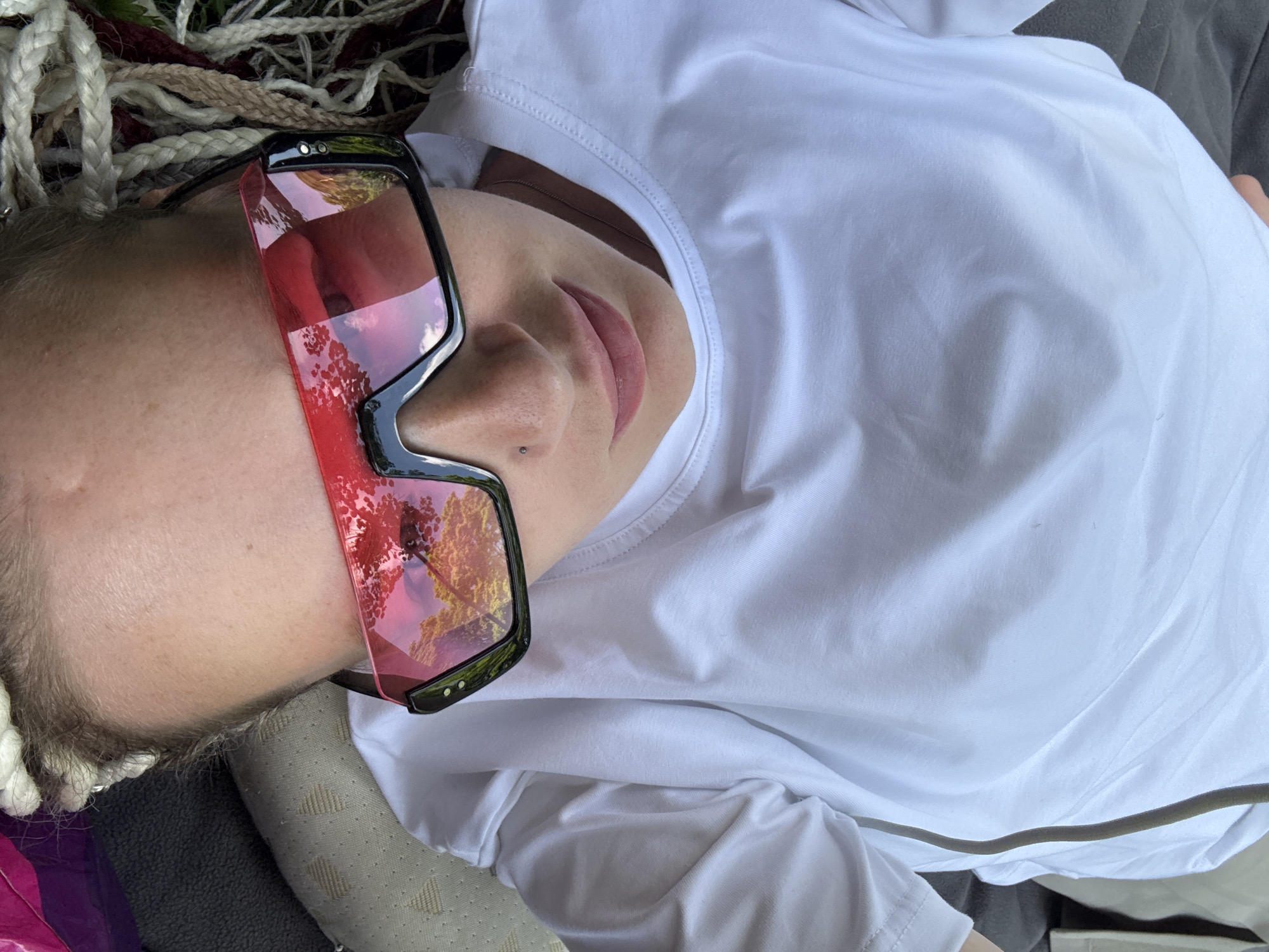 A person wearing a white long-sleeved shirt and sunglasses lies down on what appears to be a blanket or mat. The sunglasses have red lenses that reflect trees and the sky. The person's hair is in blonde braids. The image is a close-up shot, focused on the person's face and upper body. The location is unknown and cannot be determined from the image.